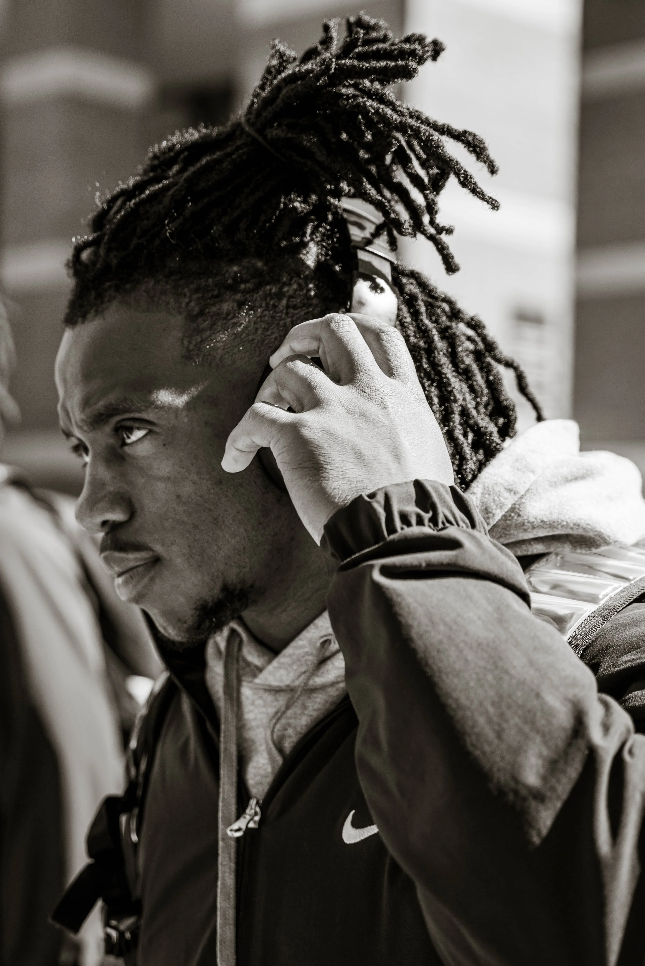 Man with dreadlocks wearing a jacket, talking on a phone, grayscale.