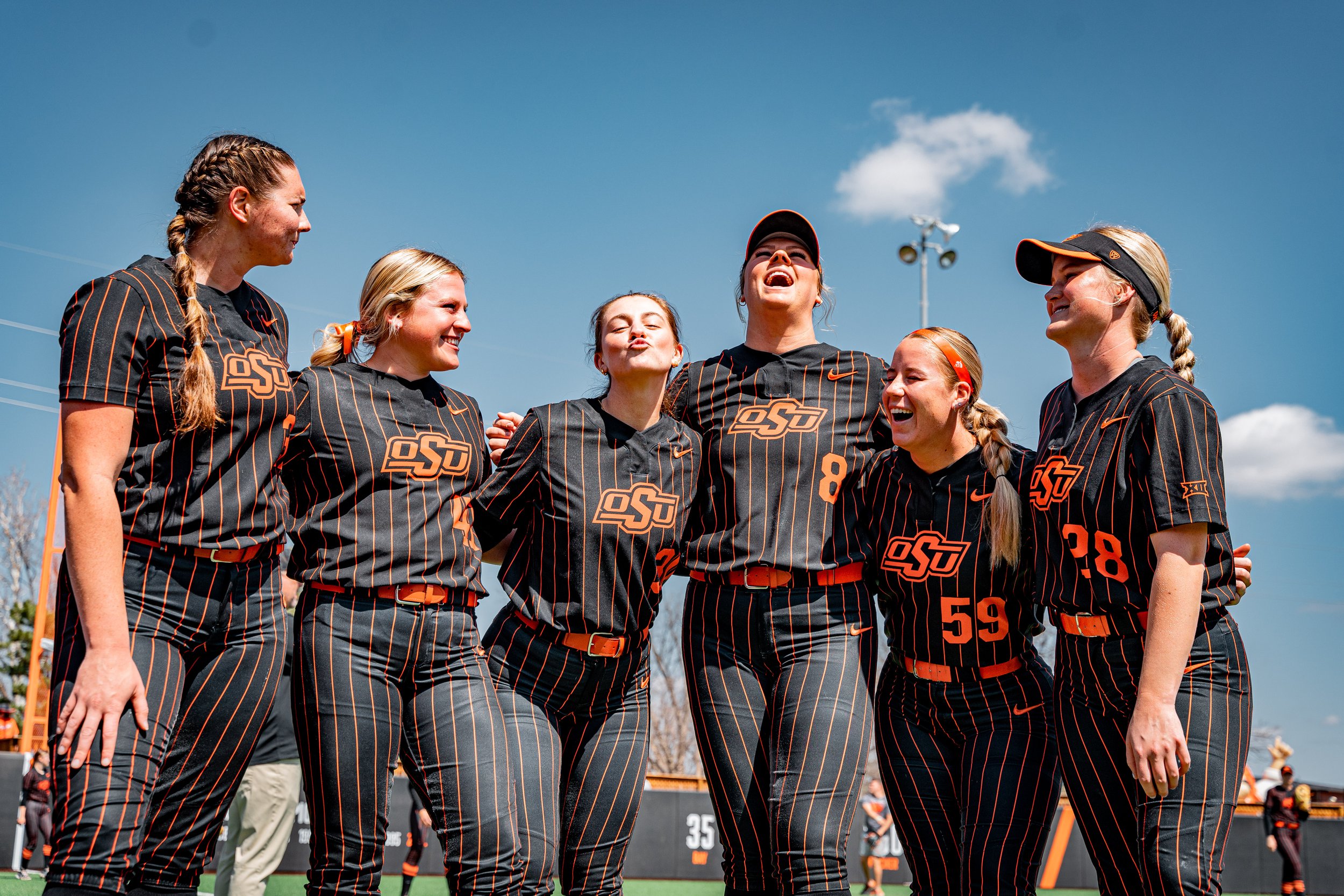 A group of six female softball players in black and orange uniforms and caps, with "OSU" logos, stand together on a field. They are smiling and appear to be celebrating or posing for a photo. The sky is clear with a few clouds visible in the backgrou