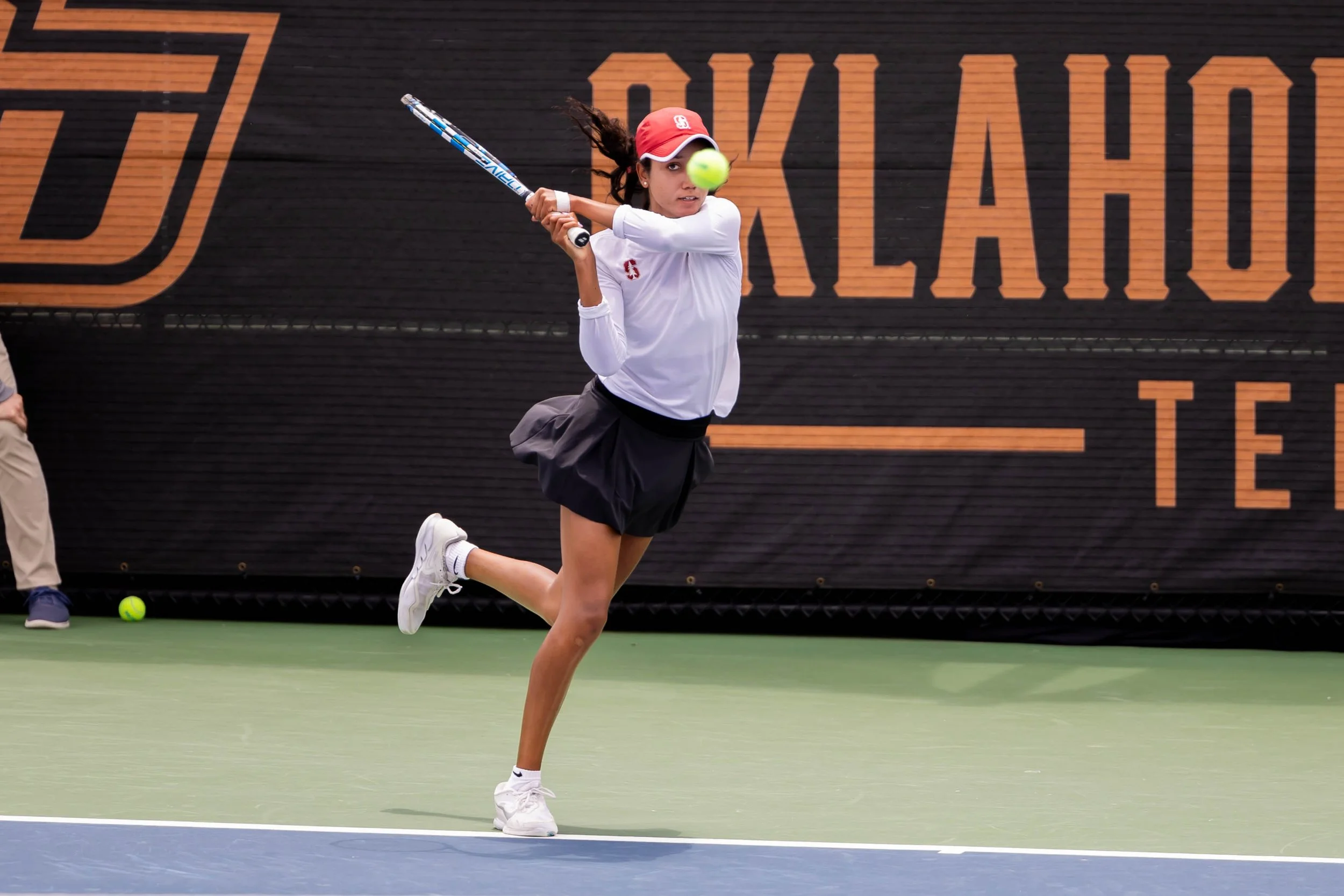 Tennis player hitting a backhand shot during a match, wearing a white shirt and black skirt, with "Oklahoma State Tennis" in the background.