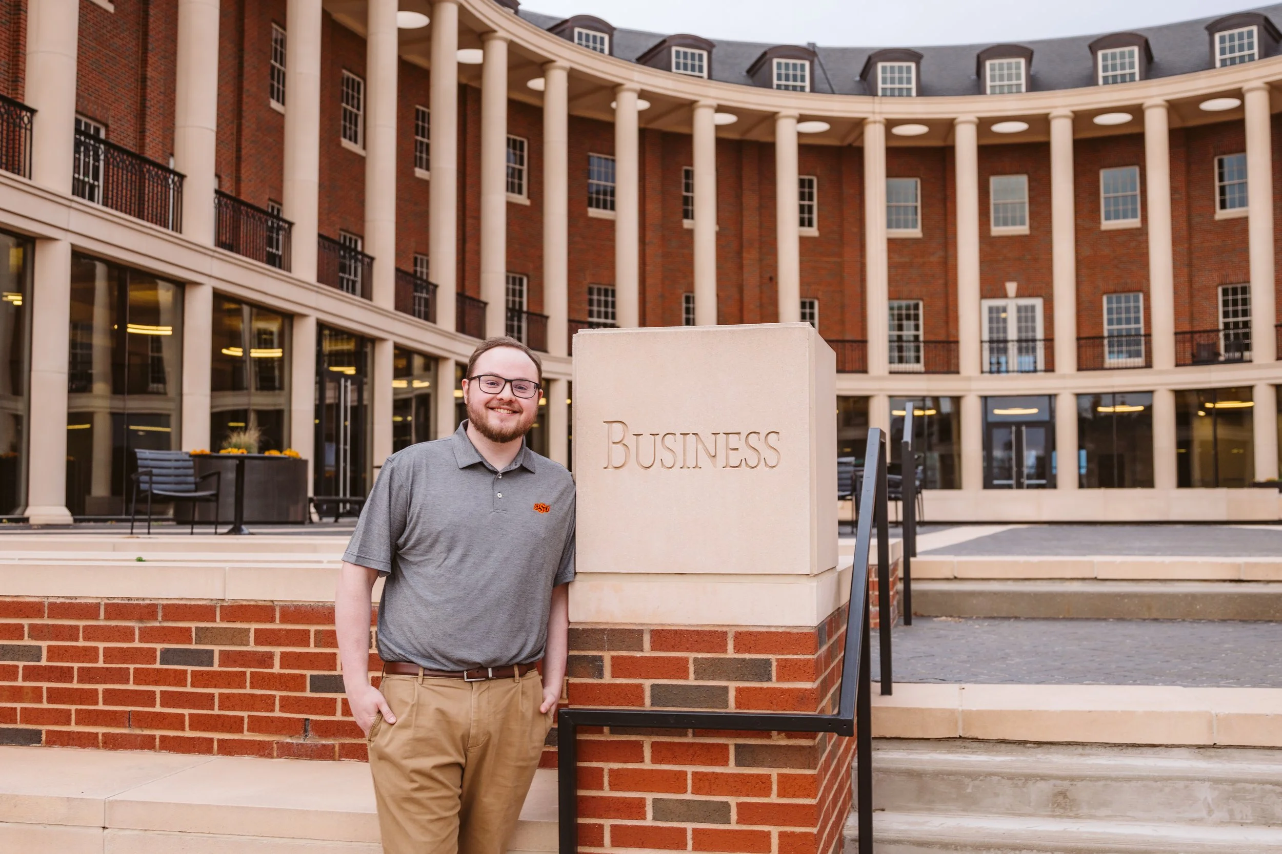 A person standing outside a building with a 'Business' sign, wearing a gray polo shirt, and surrounded by columns and brick architecture.