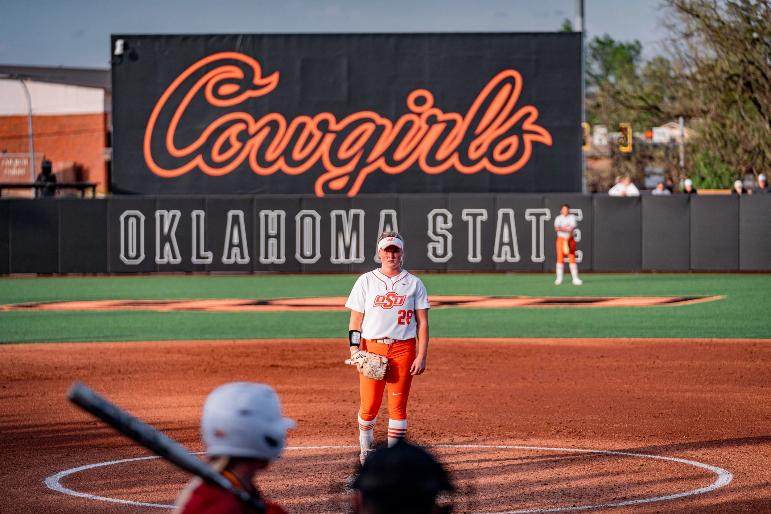 Softball player standing on the field with "Cowgirls" and "Oklahoma State" signage in the background.