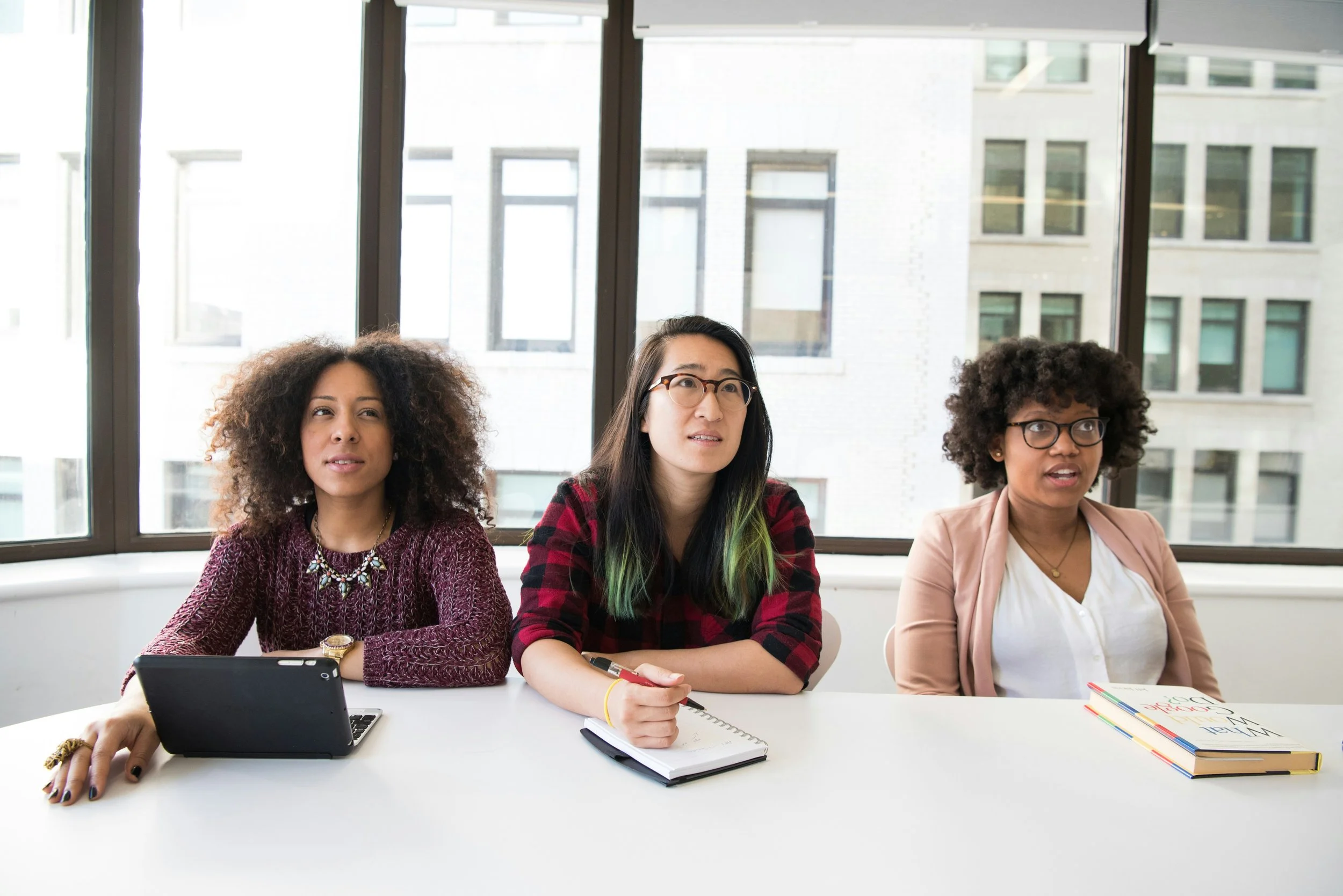 Three women sitting at a white conference table in a bright office with large windows and city buildings in the background, appearing attentive and engaged.