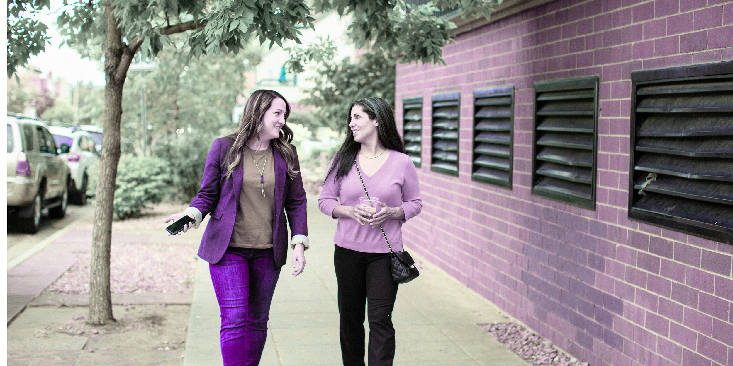 Two women walking and talking along a sidewalk next to a purple brick building with black window vents, one holding a drink and the other holding a phone.