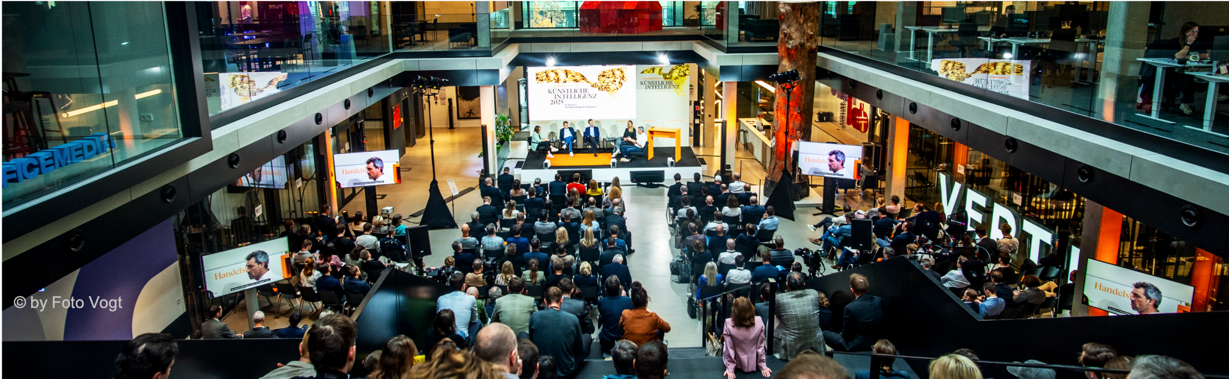 Ein Abend- oder Tagungsempfang in einem modernen zweistöckigen Veranstaltungszentrum mit vielen Zuschauern, die auf Stühlen vor einer Bühne sitzen, auf der ein Panel von fünf Sprechenden diskutiert. Mehrere große Bildschirme zeigen die Gesprächsteilnehmer. Der Veranstaltungsort ist gut beleuchtet und hat moderne Architektur mit Glaswänden und Baumskulpturen.