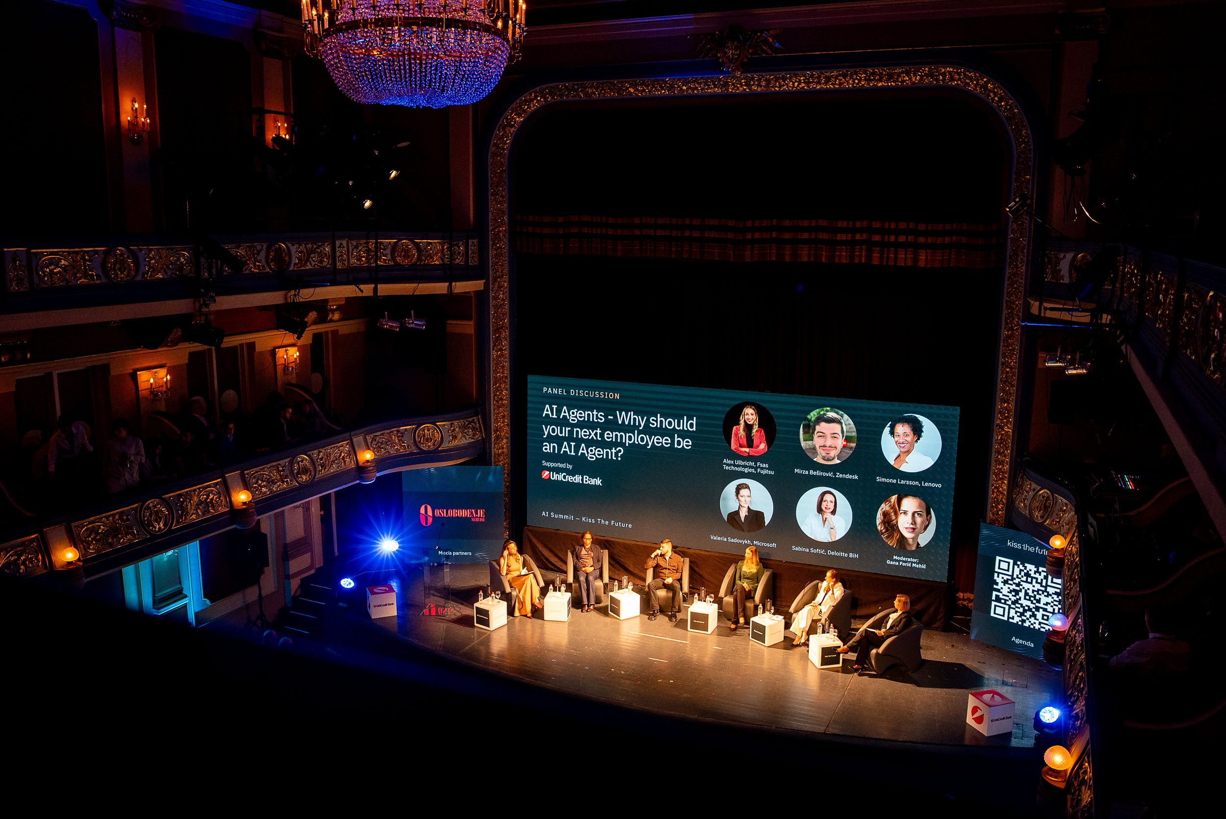 Bühne bei einer Konferenz mit Moderator*innen und Panel-Teilnehmer*innen, großer Bildschirm mit Titel 'AI Agents - Why should your next employee be an AI Agent?', eleganter Raum mit Kronleuchter und Goldverzierungen.