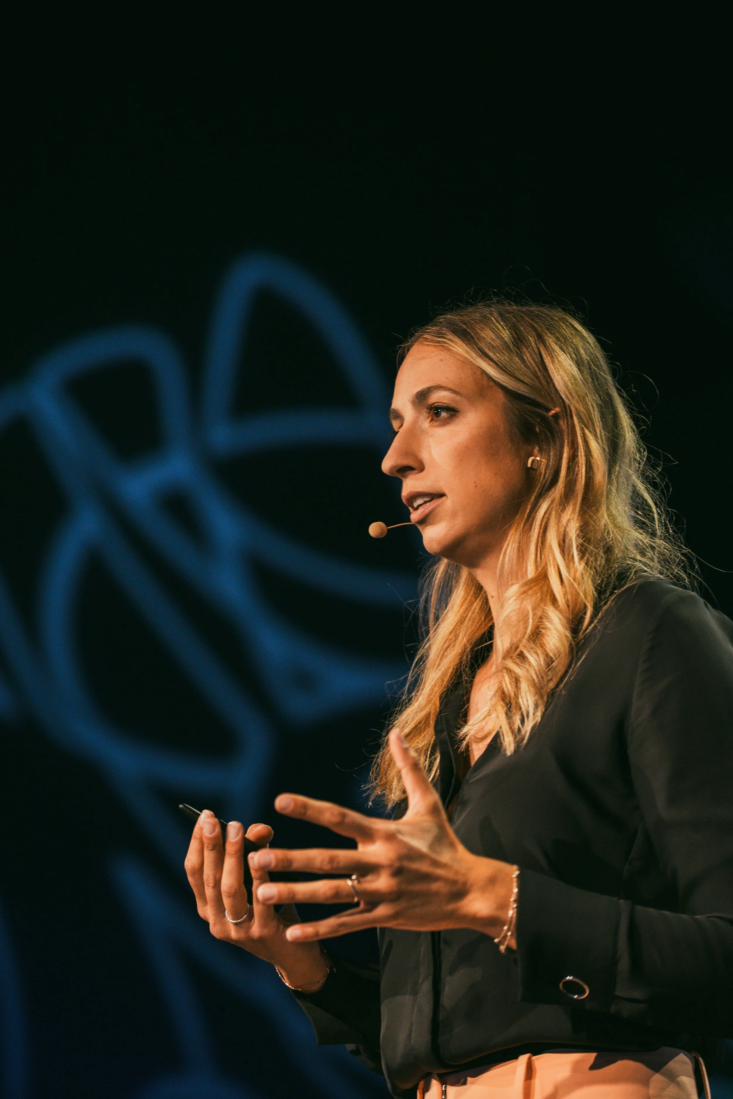 Alex Ulbricht moderiert auf einer großen Event-Bühne, Scheinwerfer. Alexandra Ulbricht hält eine Rede, sie strahlt Charme aus und begeistert das Publikum und die ZuhörerInnen. Moderatorin Alex Ulbricht leitet ein Panelgespräch zu Ethik in der KI.