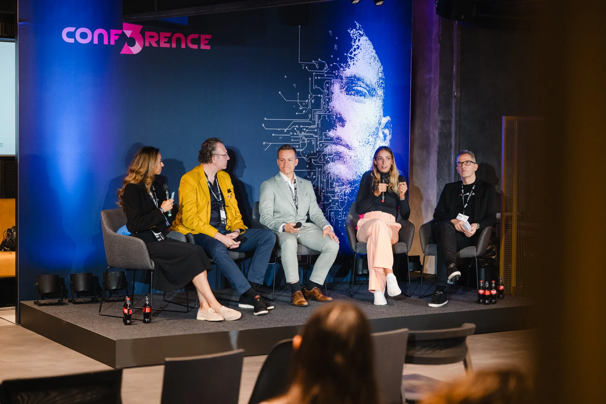 Fünf Menschen sitzen auf einer Bühne bei einer Konferenz, zwei Frauen und drei Männer, einer spricht mit einem Mikrofon, im Hintergrund ist ein digitales Gesicht und die Worte 'Conference' sichtbar.