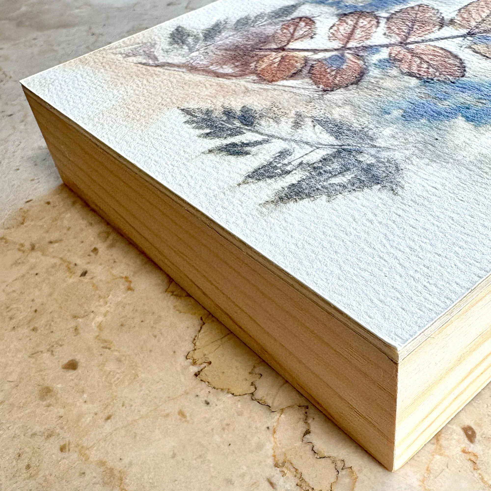 Close-up of a watercolor painting of botanical leaves on textured paper, stacked on a marble surface.