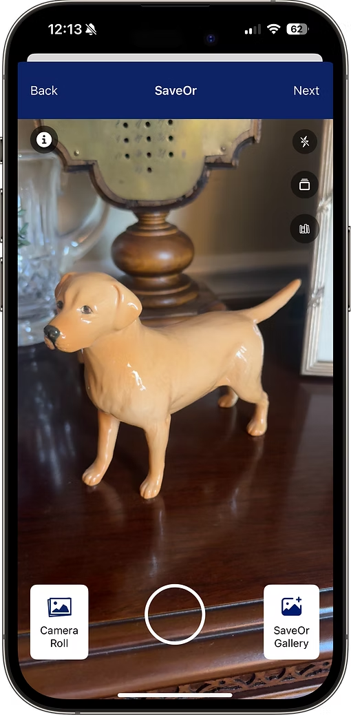 A beige ceramic dog figurine with black eyes and nose, standing on a dark wooden surface, in front of a decorative lamp and glassware.