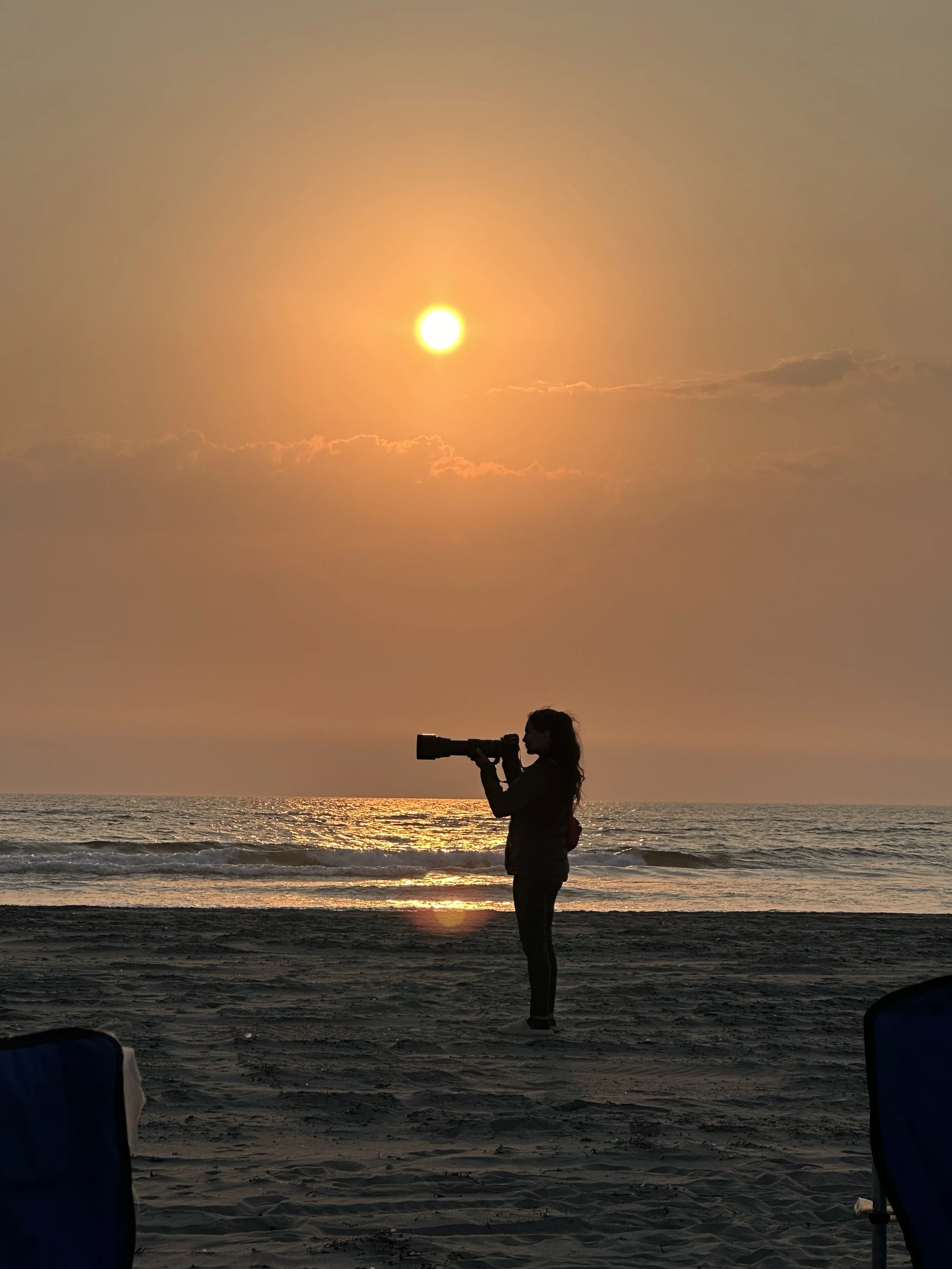 Silhouette of a woman holding a camera with a telephoto lens on a beach at sunset, with the sun low in the sky and ocean waves in the background.