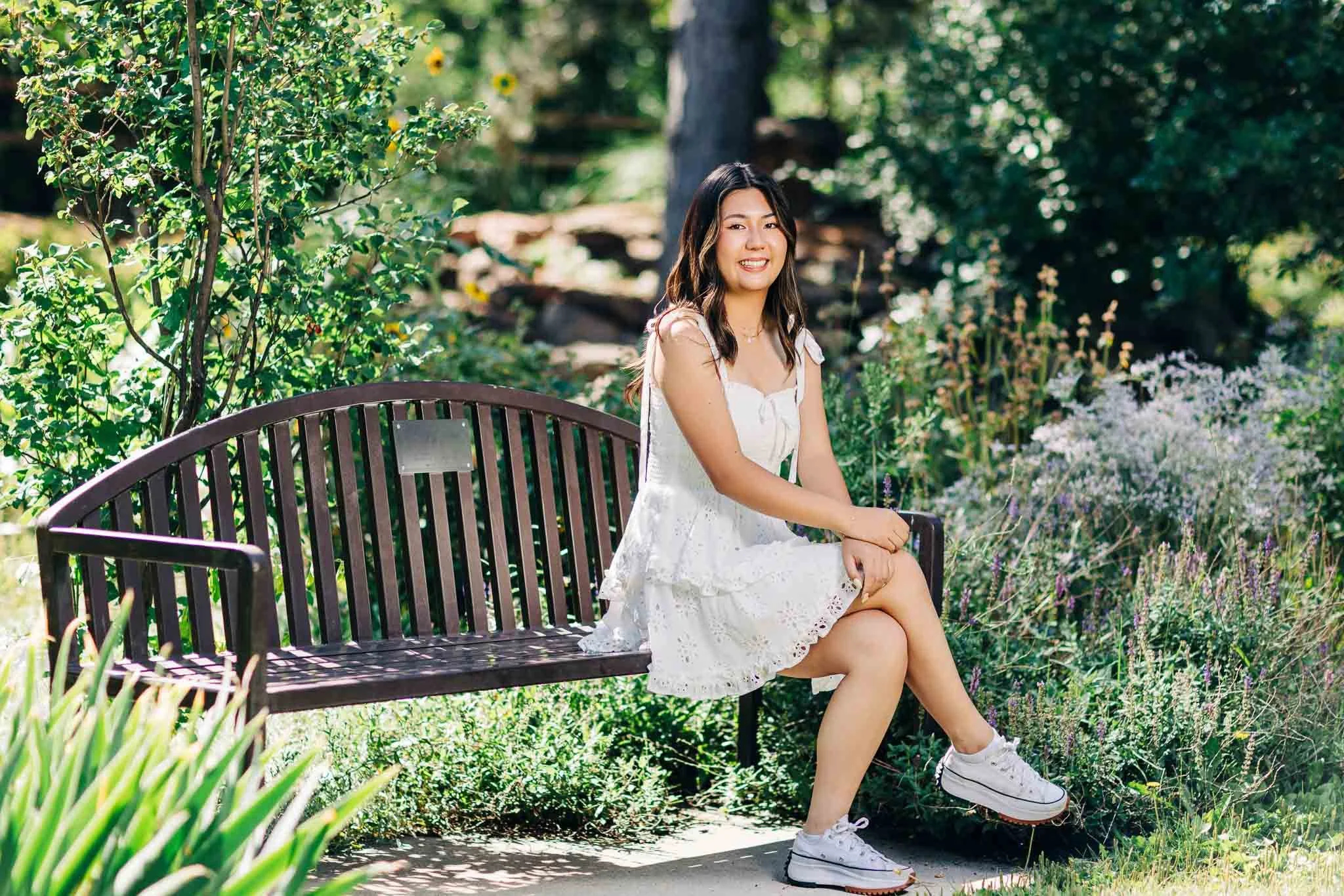 A young woman sitting on a black metal park bench in a garden with green plants and flowers, smiling at the camera, wearing a white dress and sneakers.