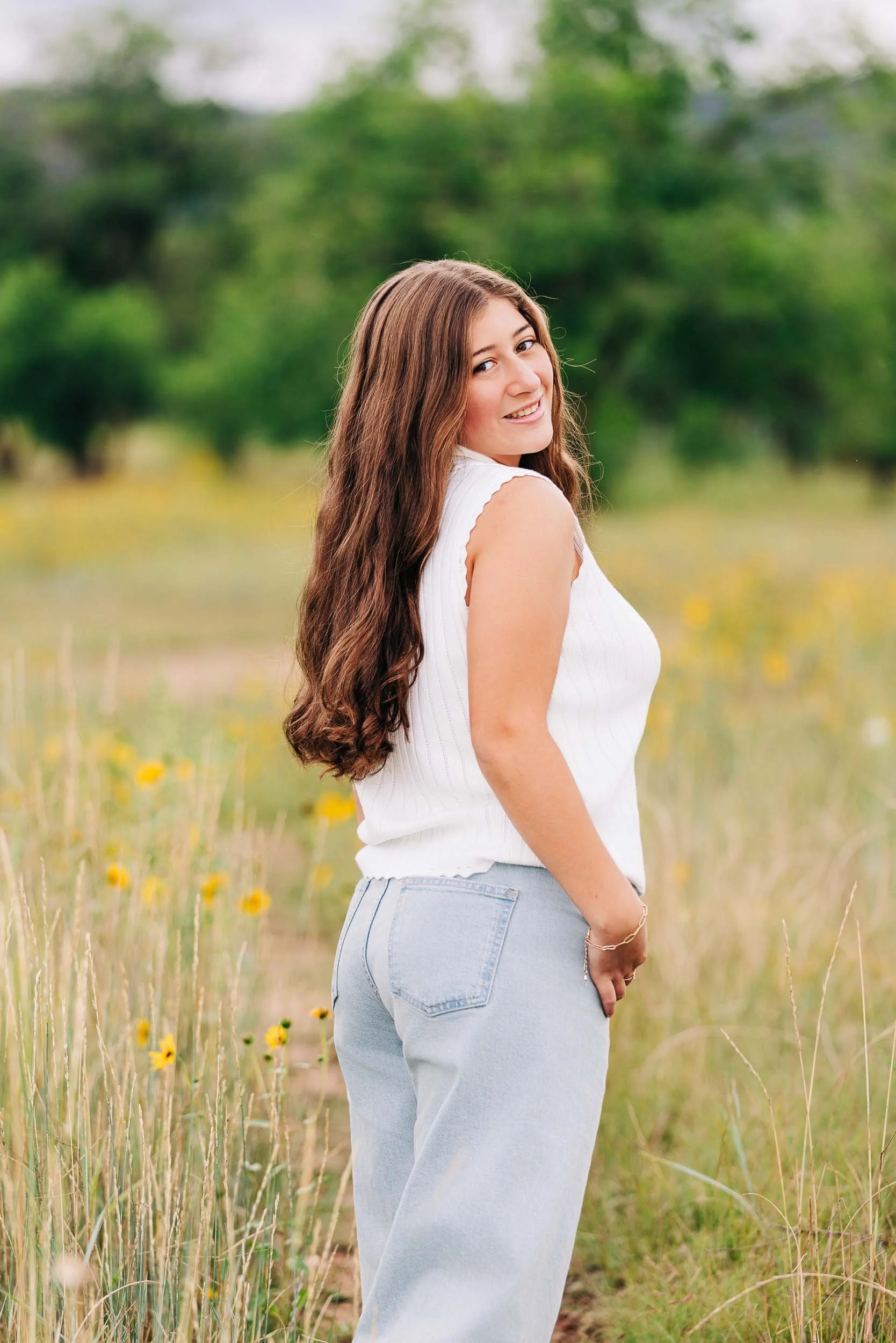 A high school girl with long, wavy brown hair smiling in a field of yellow flowers and tall grass, wearing a white sleeveless top and light blue jeans.