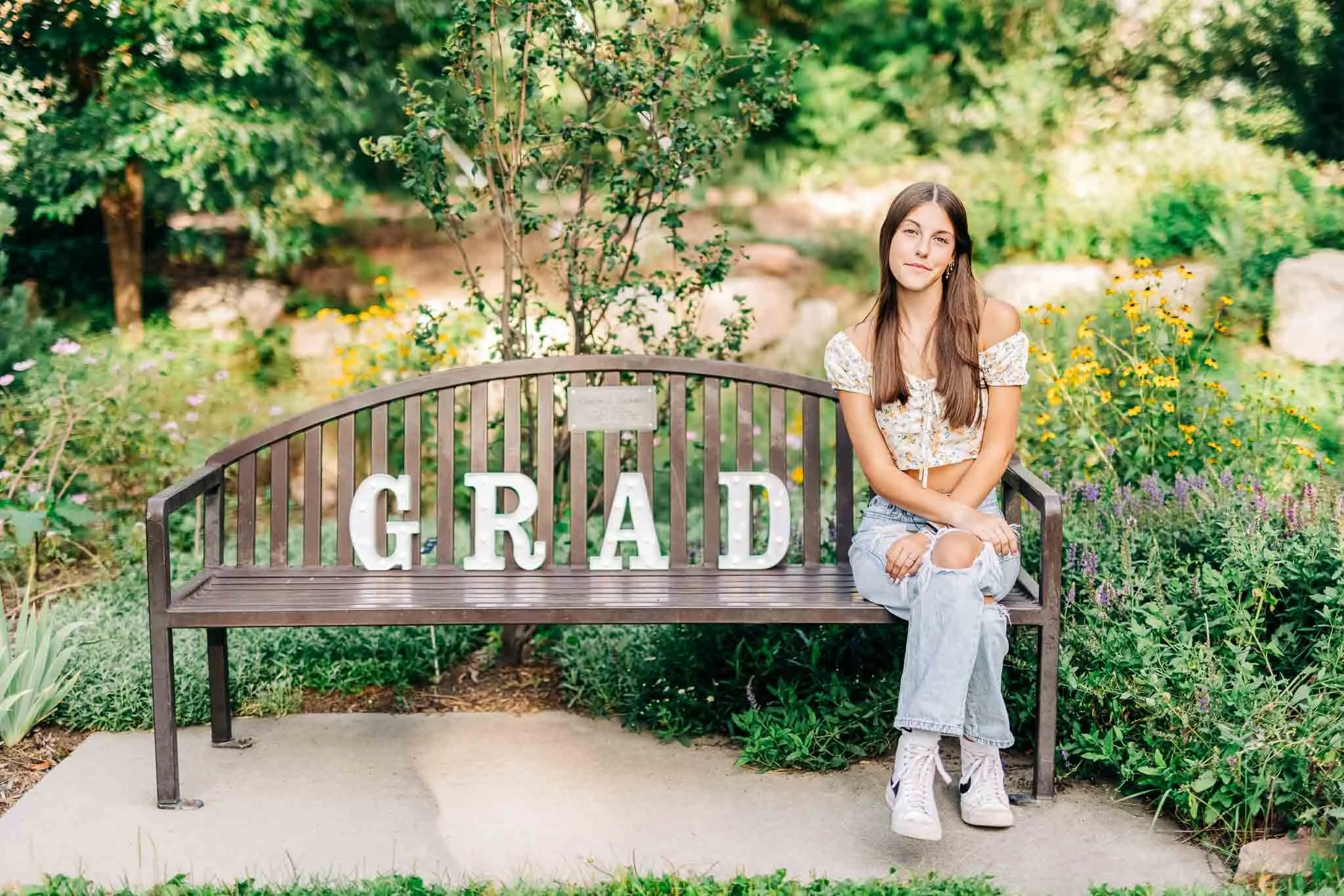 A young woman sitting on a bench in a garden, with large letter signs spelling 'GRAD' on the bench behind her, surrounded by colorful flowers and greenery.