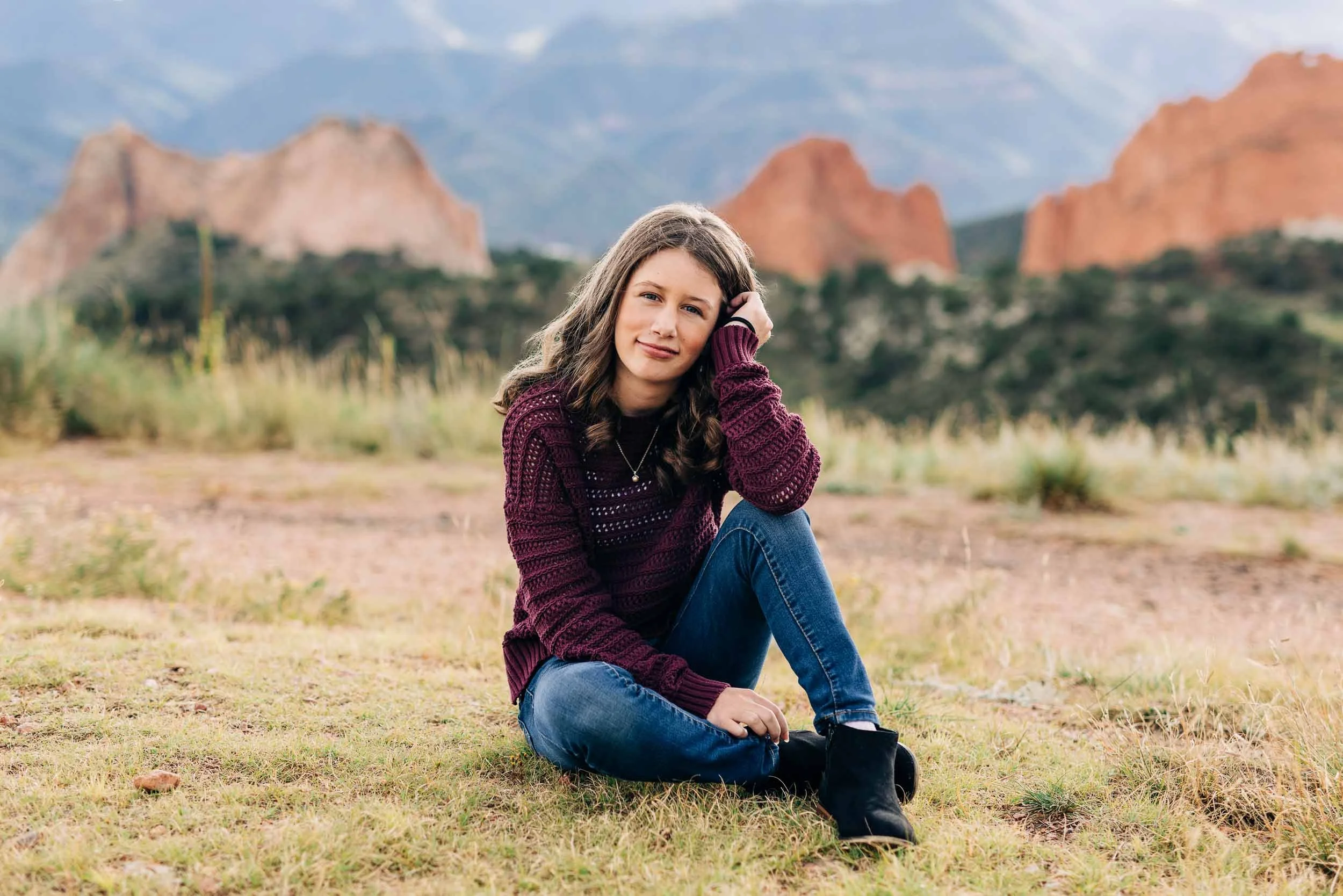 A teen girl with brown hair sitting on the grass in a natural landscape with rocks and mountains in the background, wearing a maroon sweater, blue jeans, and black boots.