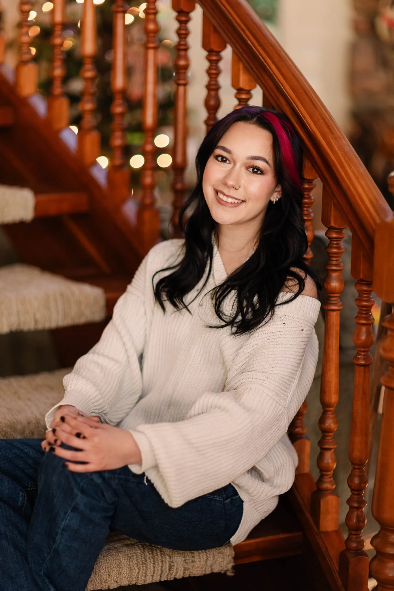 A young woman with long black hair with pink streaks, sitting on carpeted stairs in a warmly lit home, smiling at the camera, wearing a cream off-the-shoulder sweater and dark jeans.