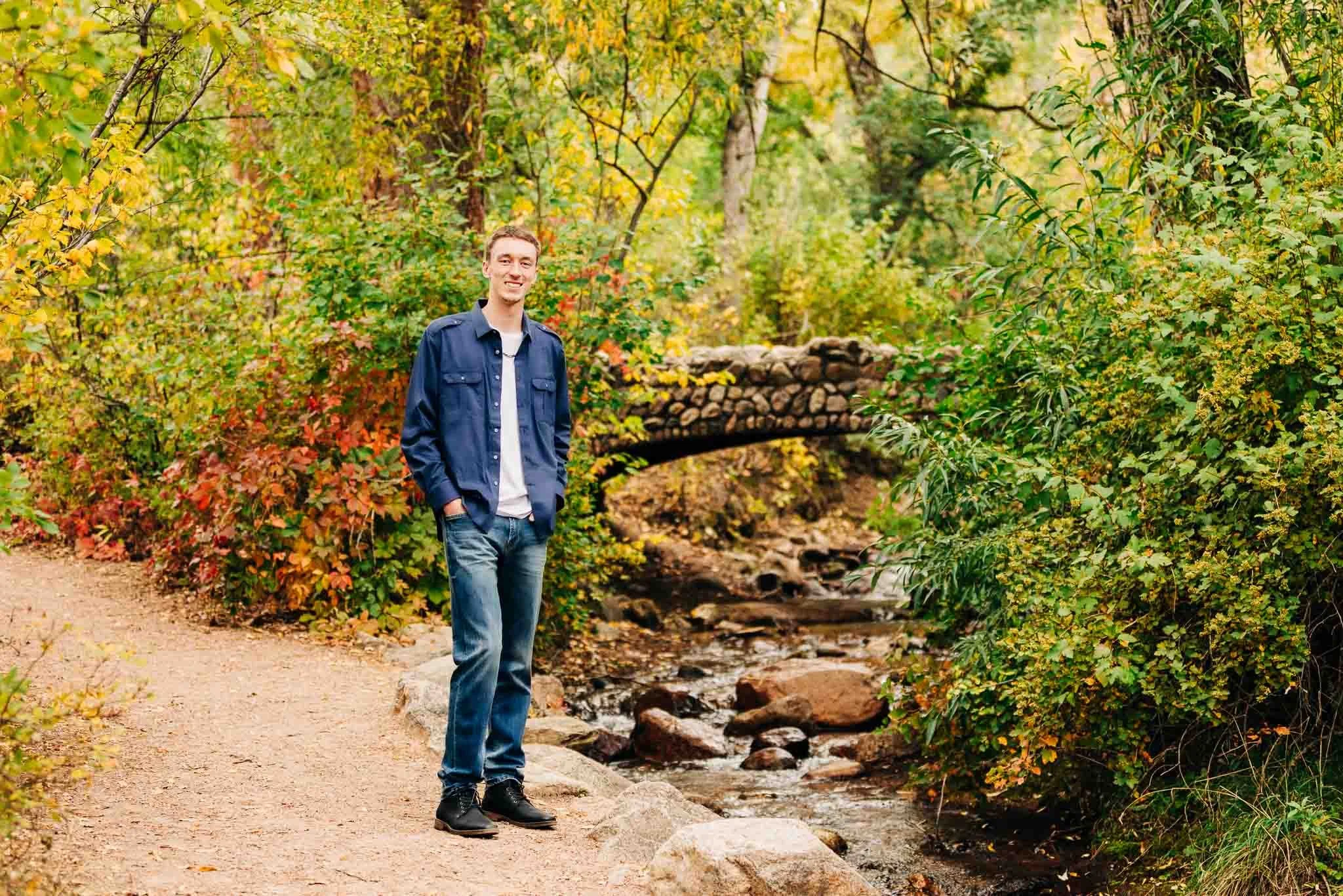 A young man with light skin and brown hair, wearing a blue jacket, white T-shirt, and jeans, standing on a dirt trail beside a small creek surrounded by colorful autumn foliage, with a small stone bridge in the background.