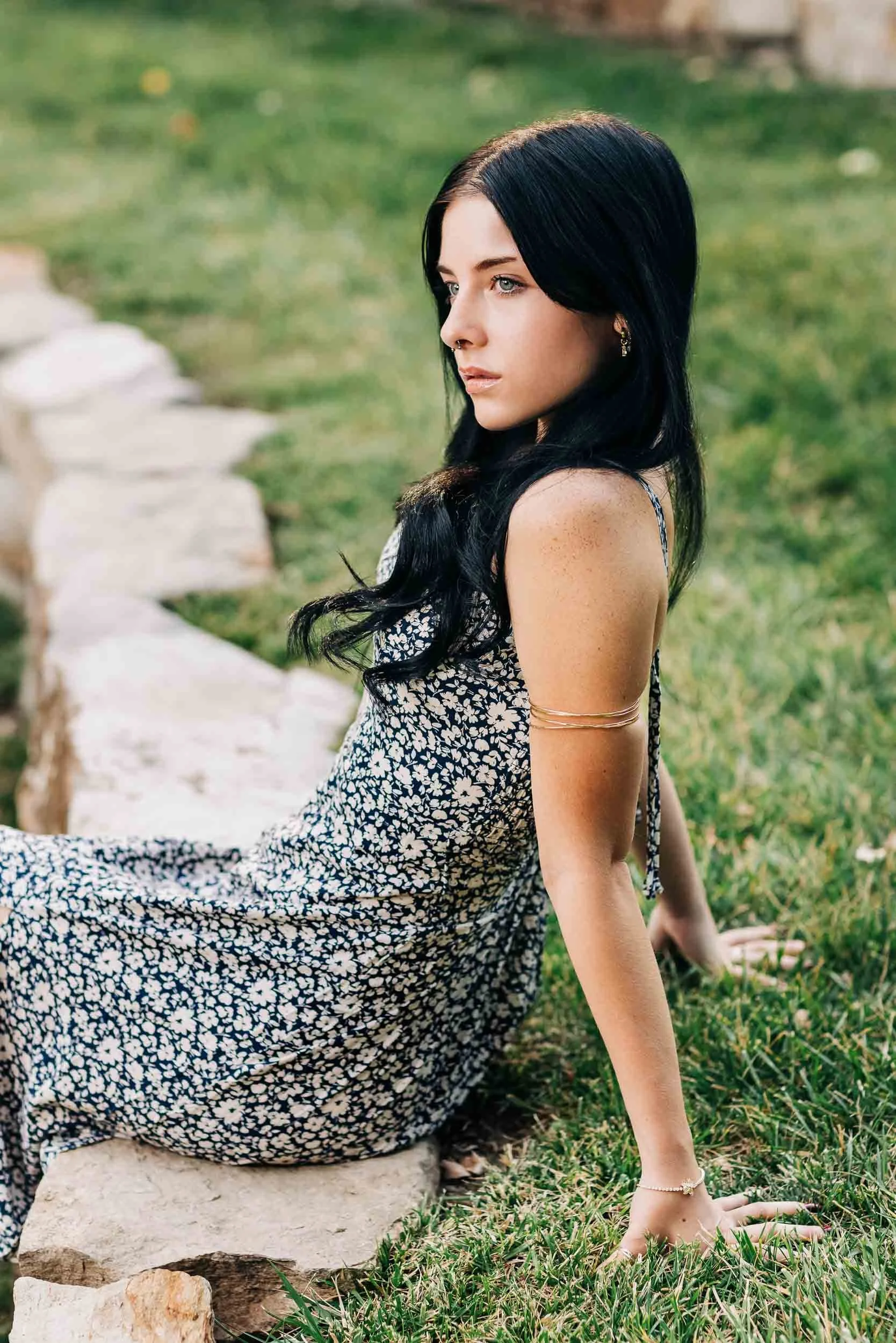 A high school senior girl with black hair, wearing a floral dress, is sitting outdoors on grass, leaning back with one hand on the ground and the other resting on her knee, looking into the distance.