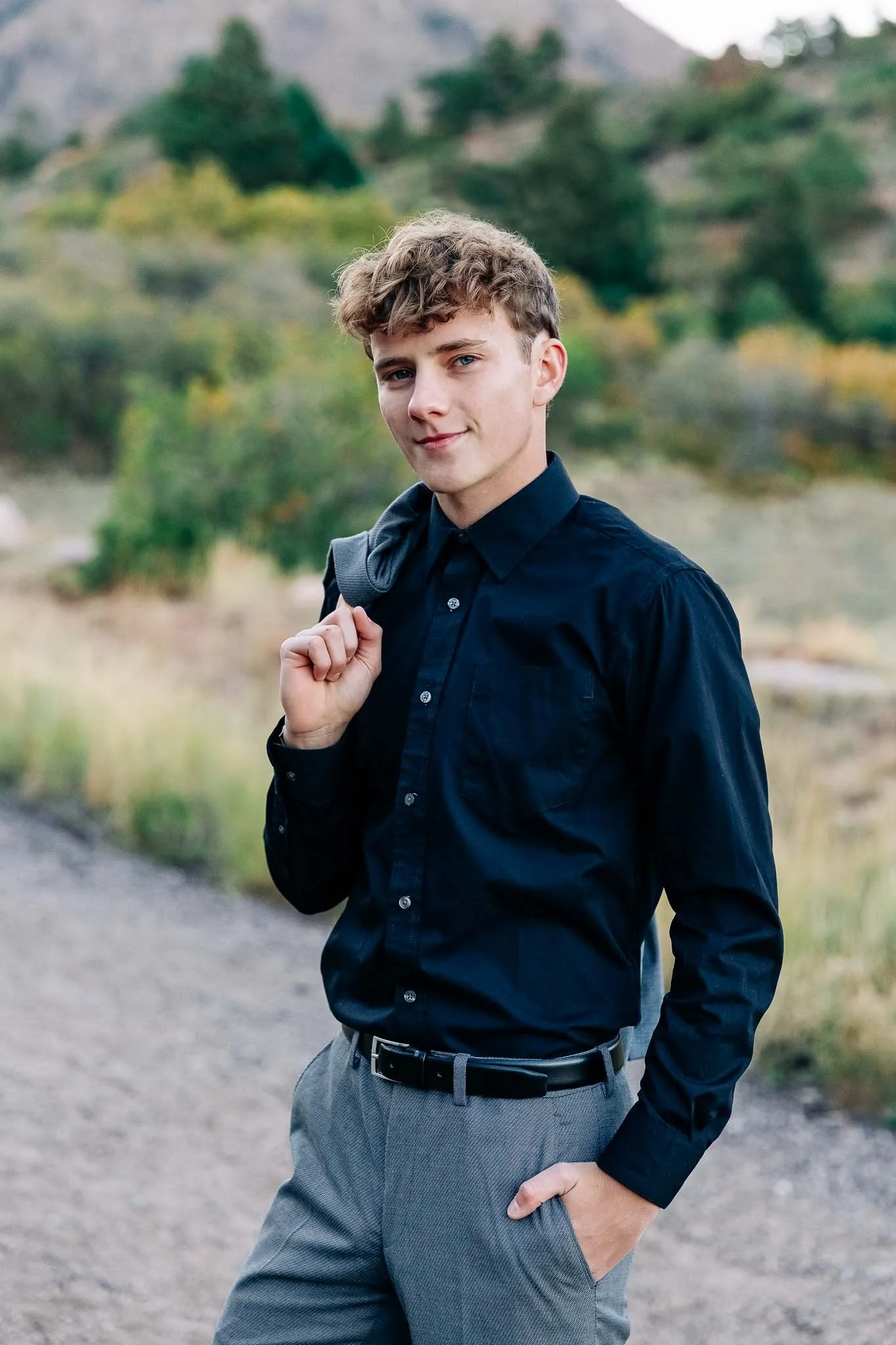 A young man standing outdoors on a dirt path, wearing a dark blue button-up shirt and gray pants, holding a gray jacket over his shoulder, with a scenic background of trees and hills.