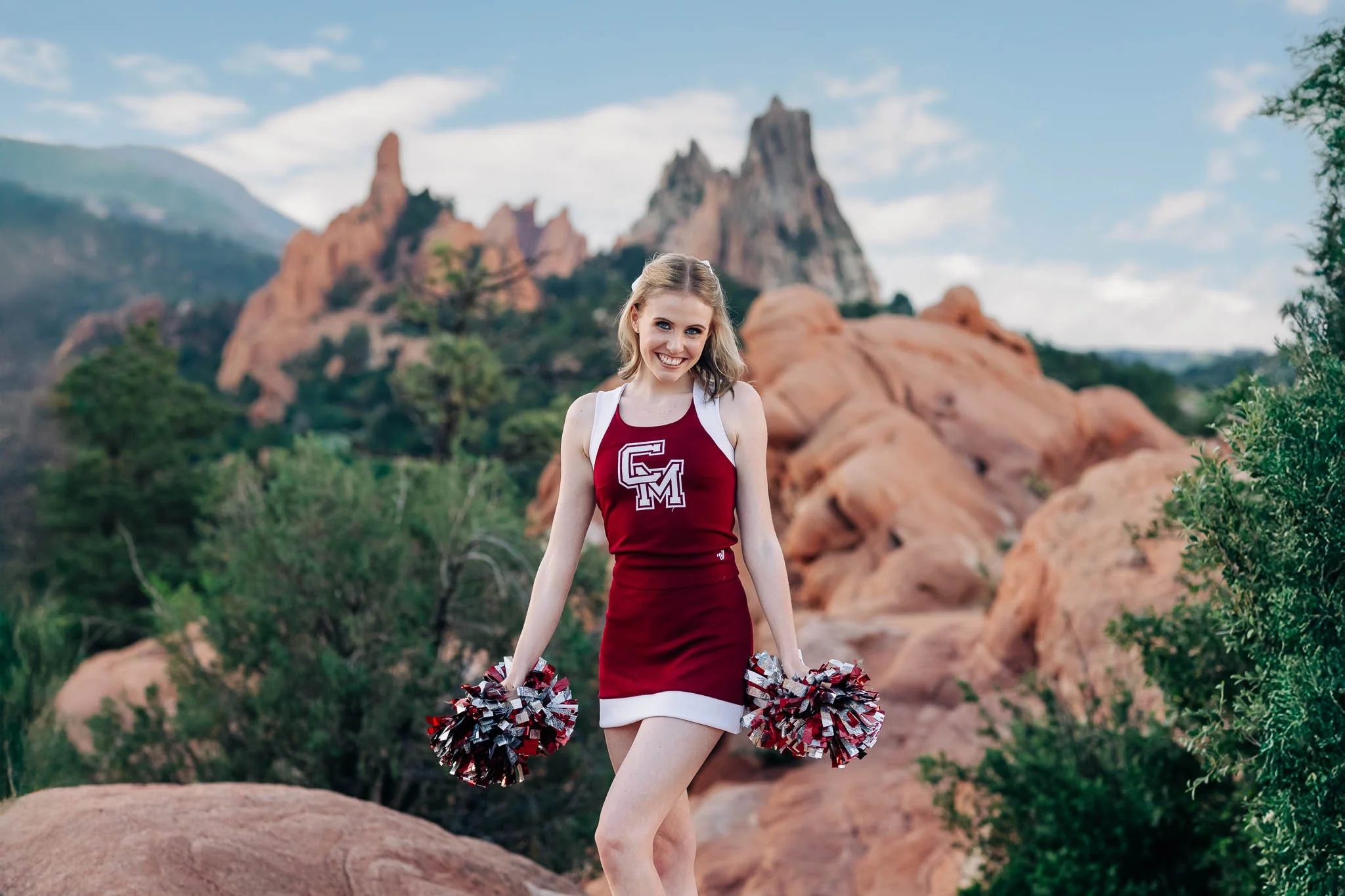 A smiling cheerleader with blonde hair in a red and white uniform holding pom-poms, standing outdoors with rocks and trees in the background under a partly cloudy sky.