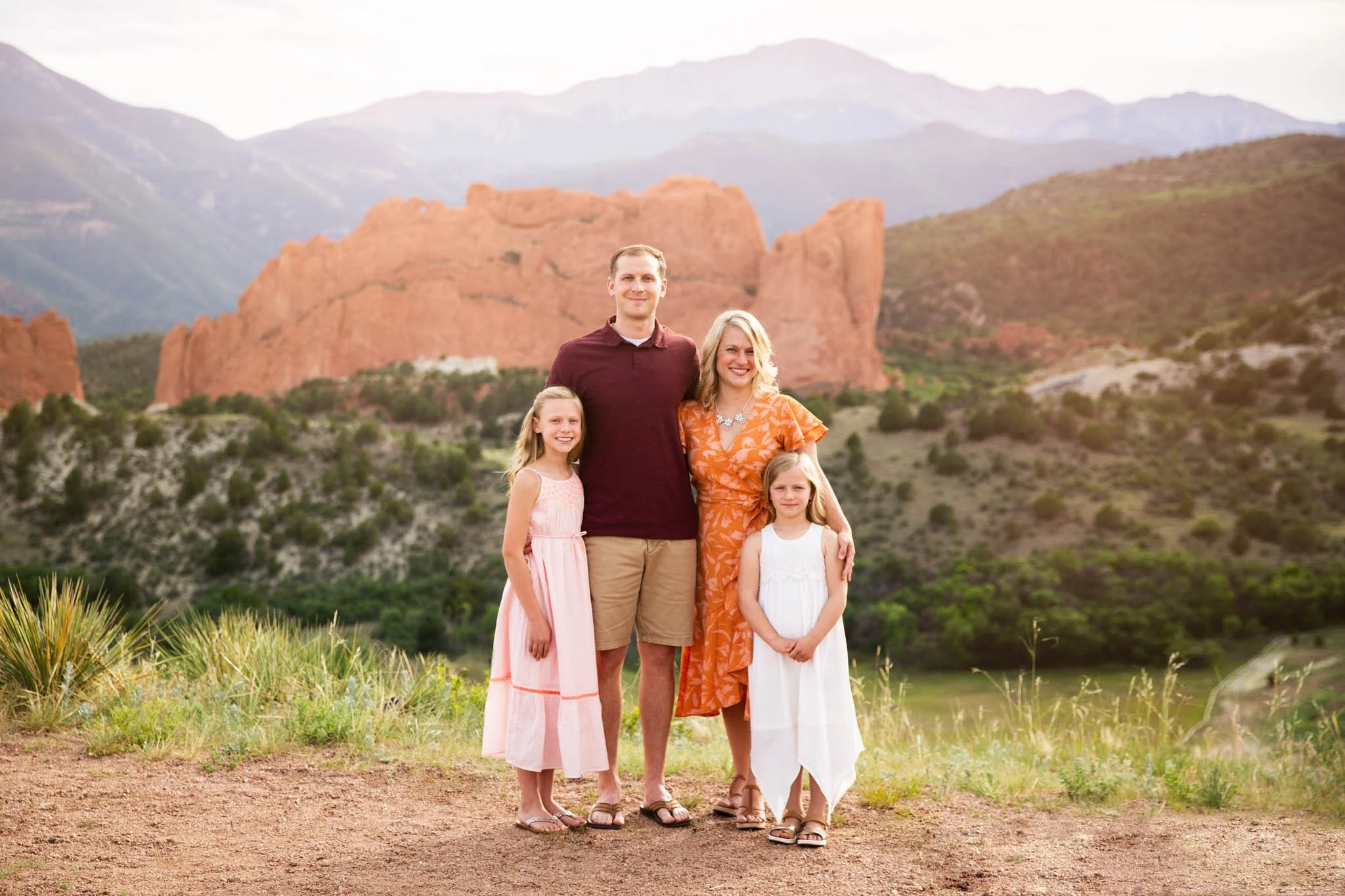 A family of five standing outdoors with red rock formations and mountains in the background, during sunset, smiling at the camera.
