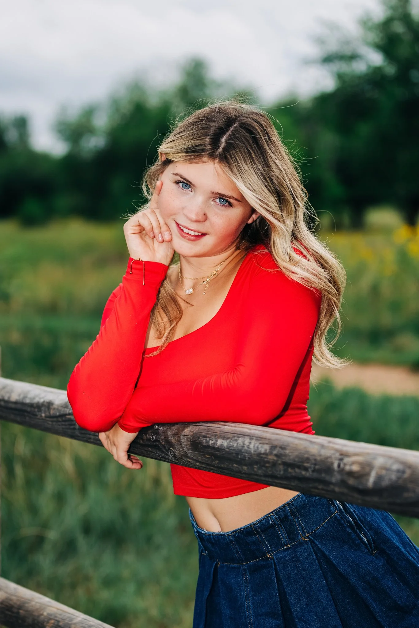 A high school junior with long blonde hair and blue eyes, wearing a red long-sleeve top and blue jeans, leaning on a wooden fence in an outdoor setting with green trees in the background.