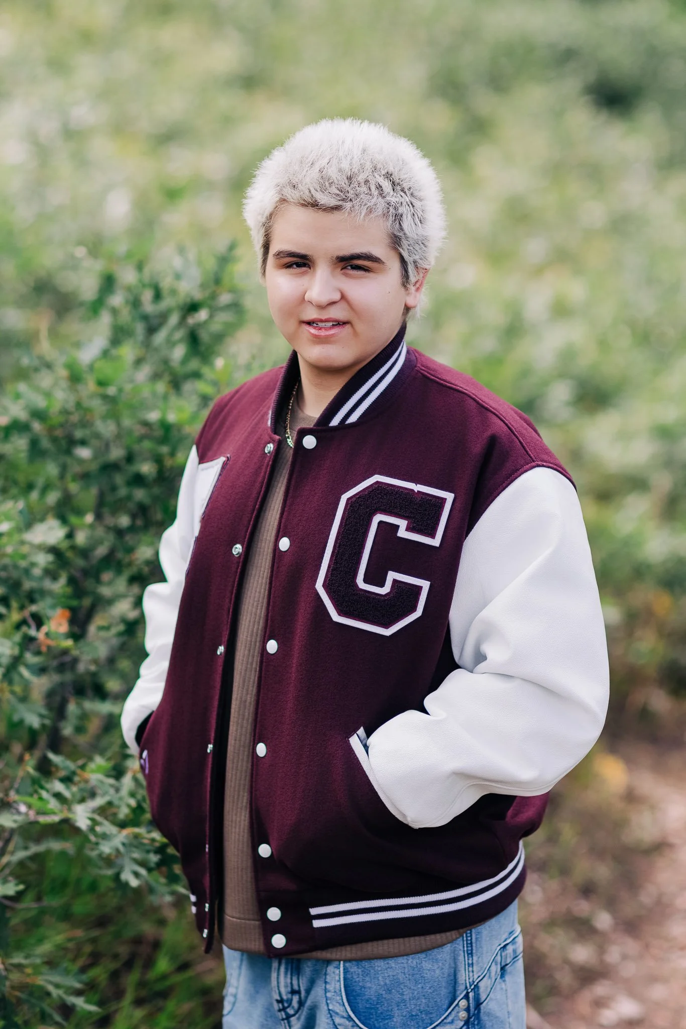 A high school senior guy with short, bleached hair and light complexion wearing a maroon and white Cheyenne Mountain High School varsity jacket, standing outdoors in a natural, leafy environment.