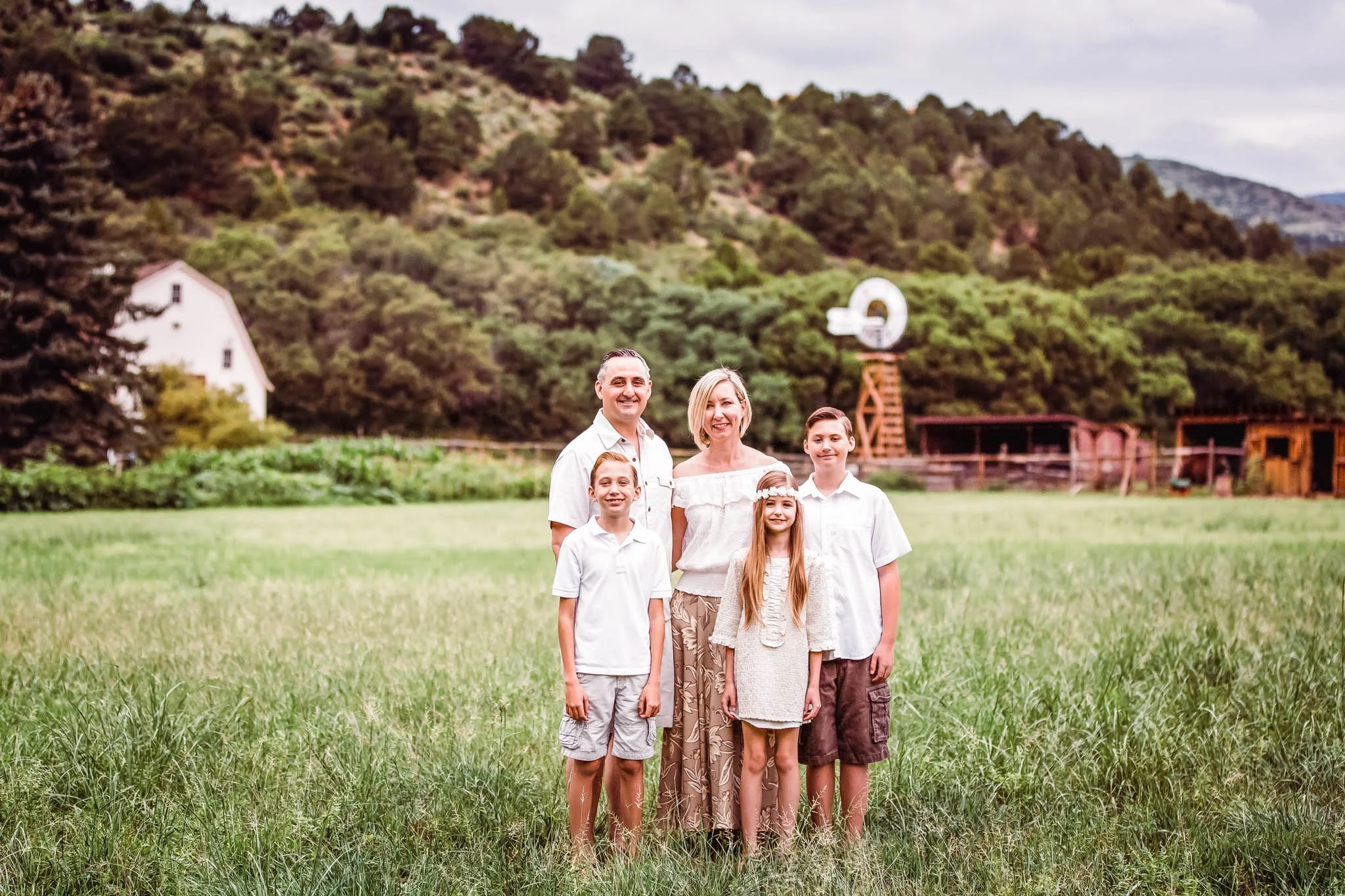Family of five standing in a grassy field with trees, hills, a white house, and a windmill in the background.