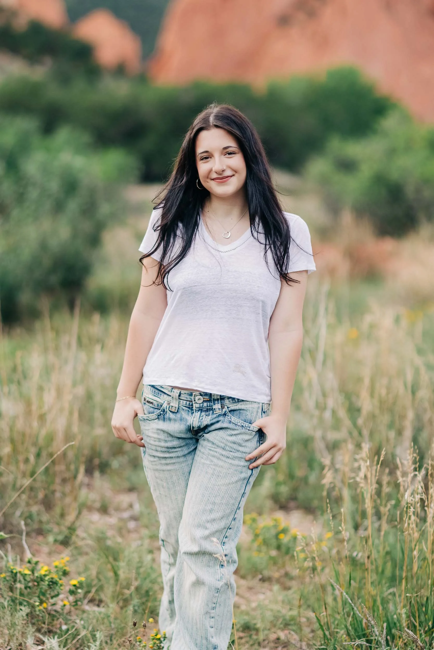 A high school senior girl with long dark hair standing outdoors in a grassy field with trees and large rocks in the background. She is smiling and wearing a light-colored T-shirt and jeans.