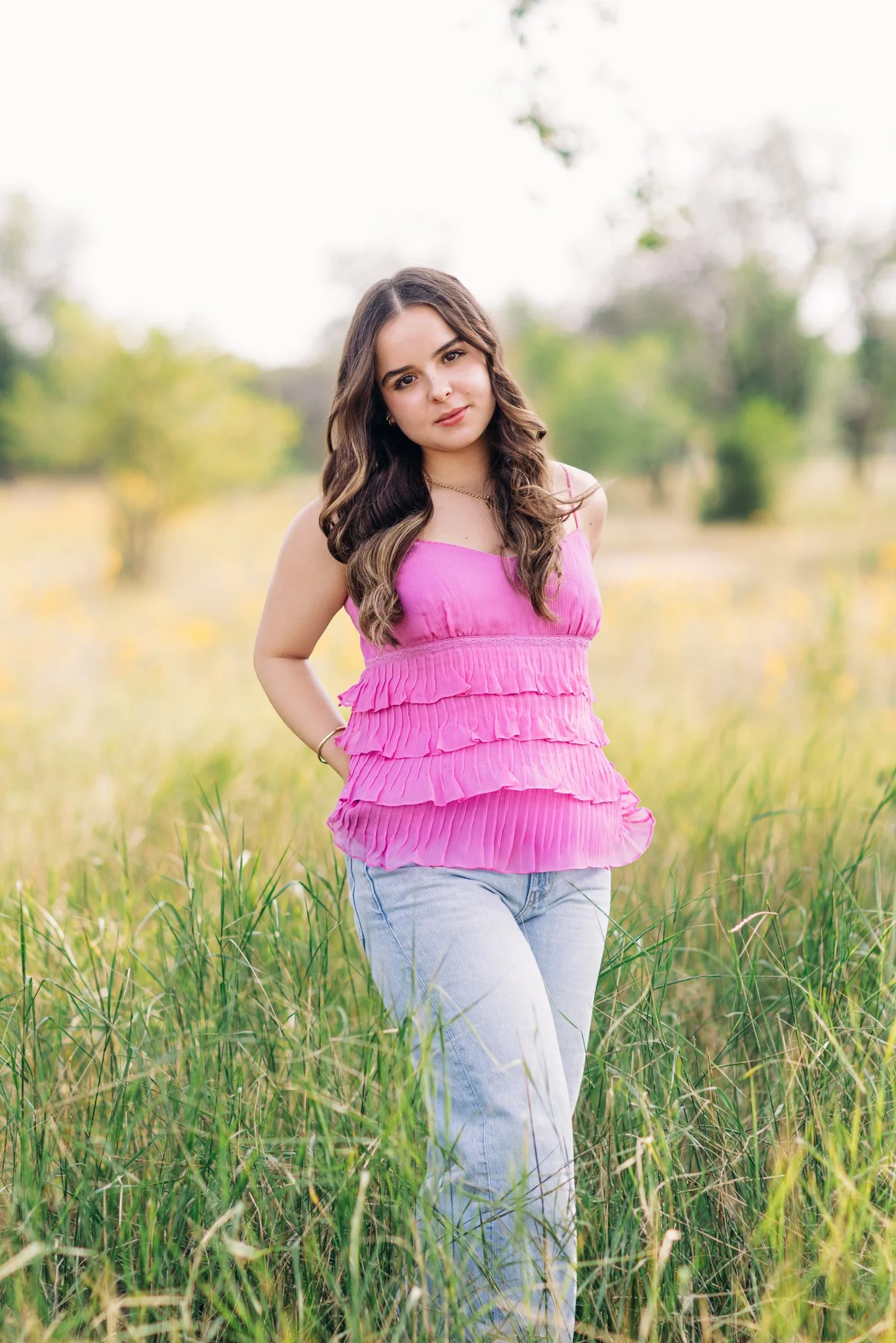 A high school senior with long wavy brown hair wearing a pink ruffled top and light blue jeans standing in a grassy field with trees in the background during daytime.