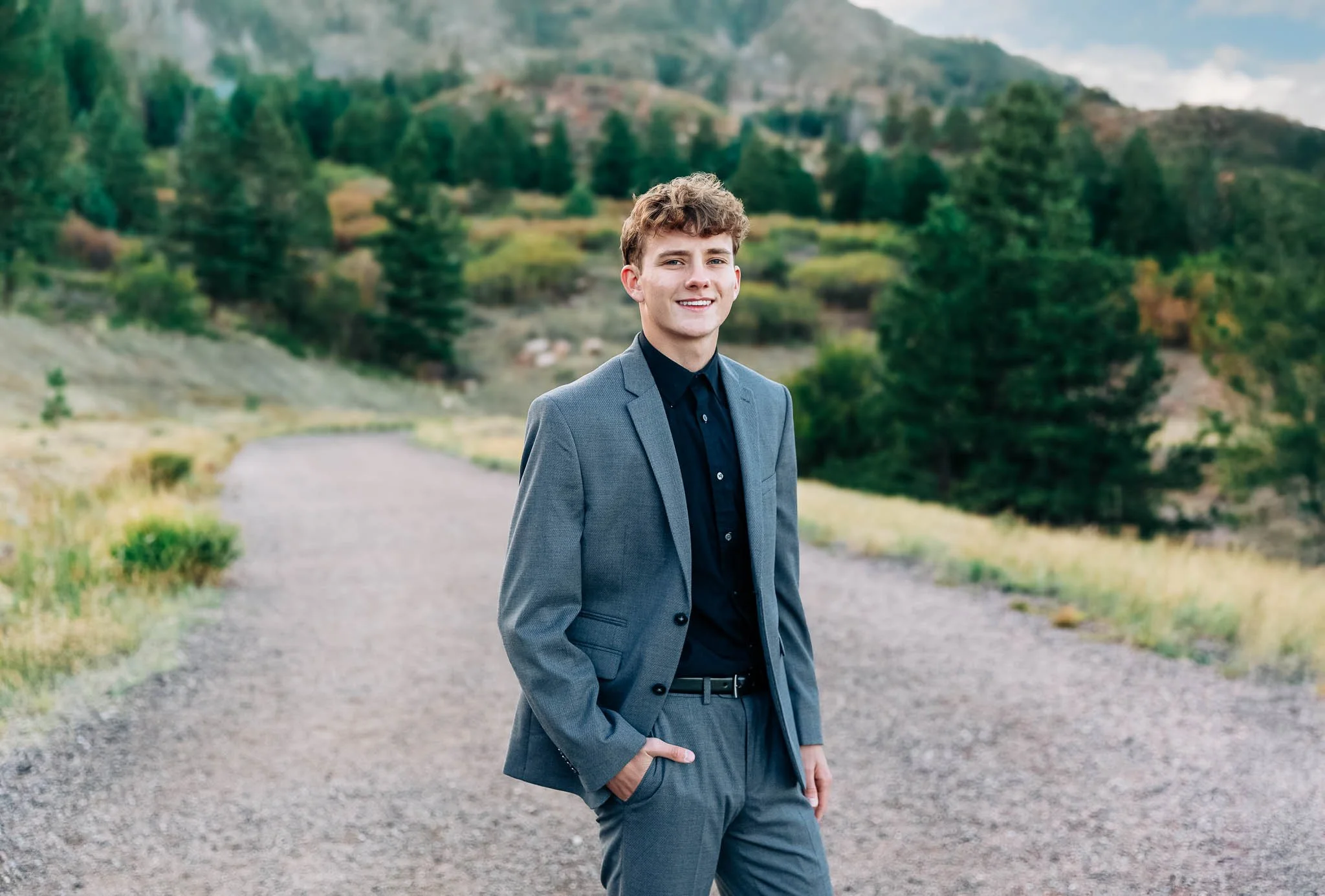 A young man wearing a grey suit and black shirt standing outdoors on a gravel path with a scenic background of trees and hills.