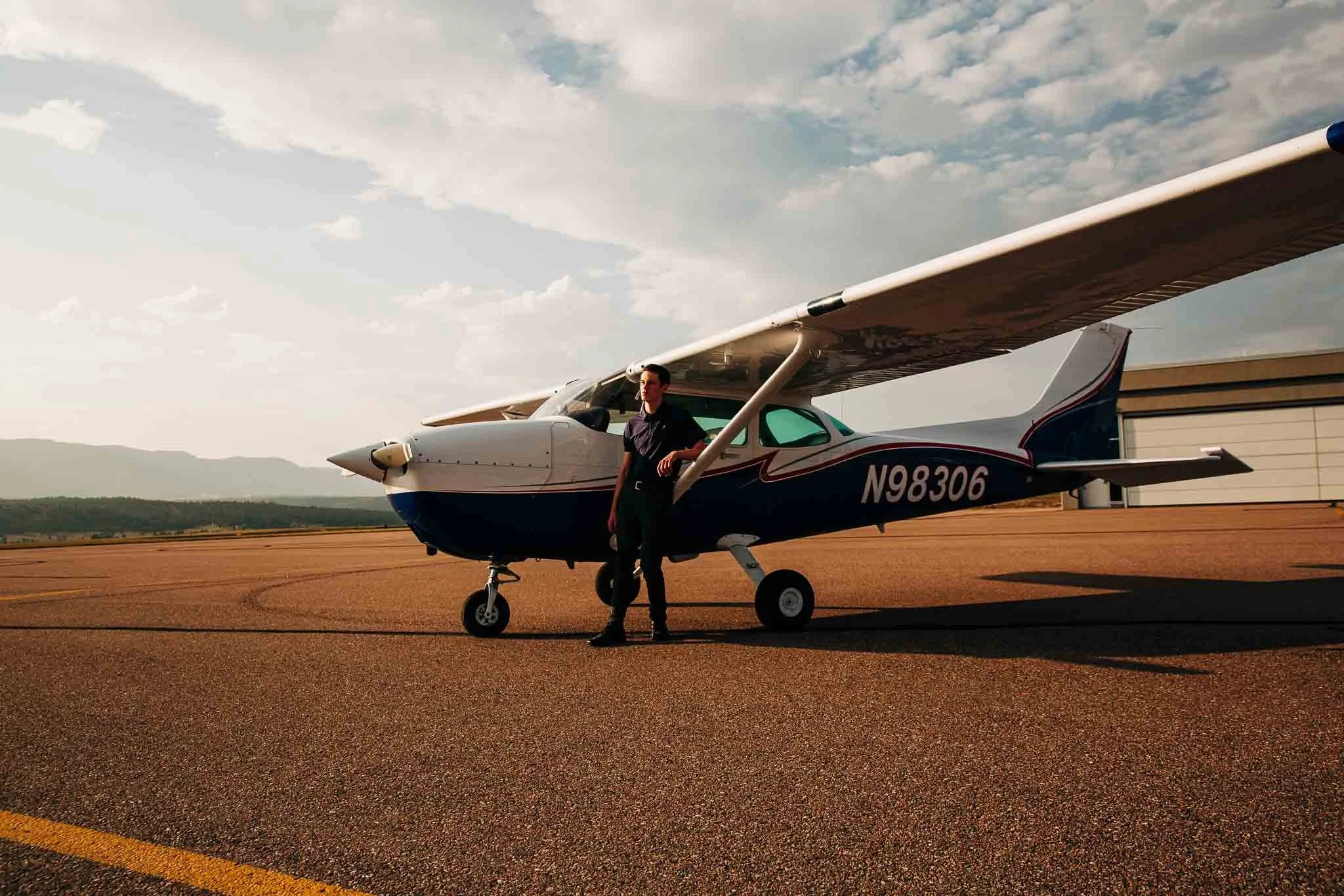 Man standing beside small single-engine aircraft on airfield during sunset with mountains in the background.