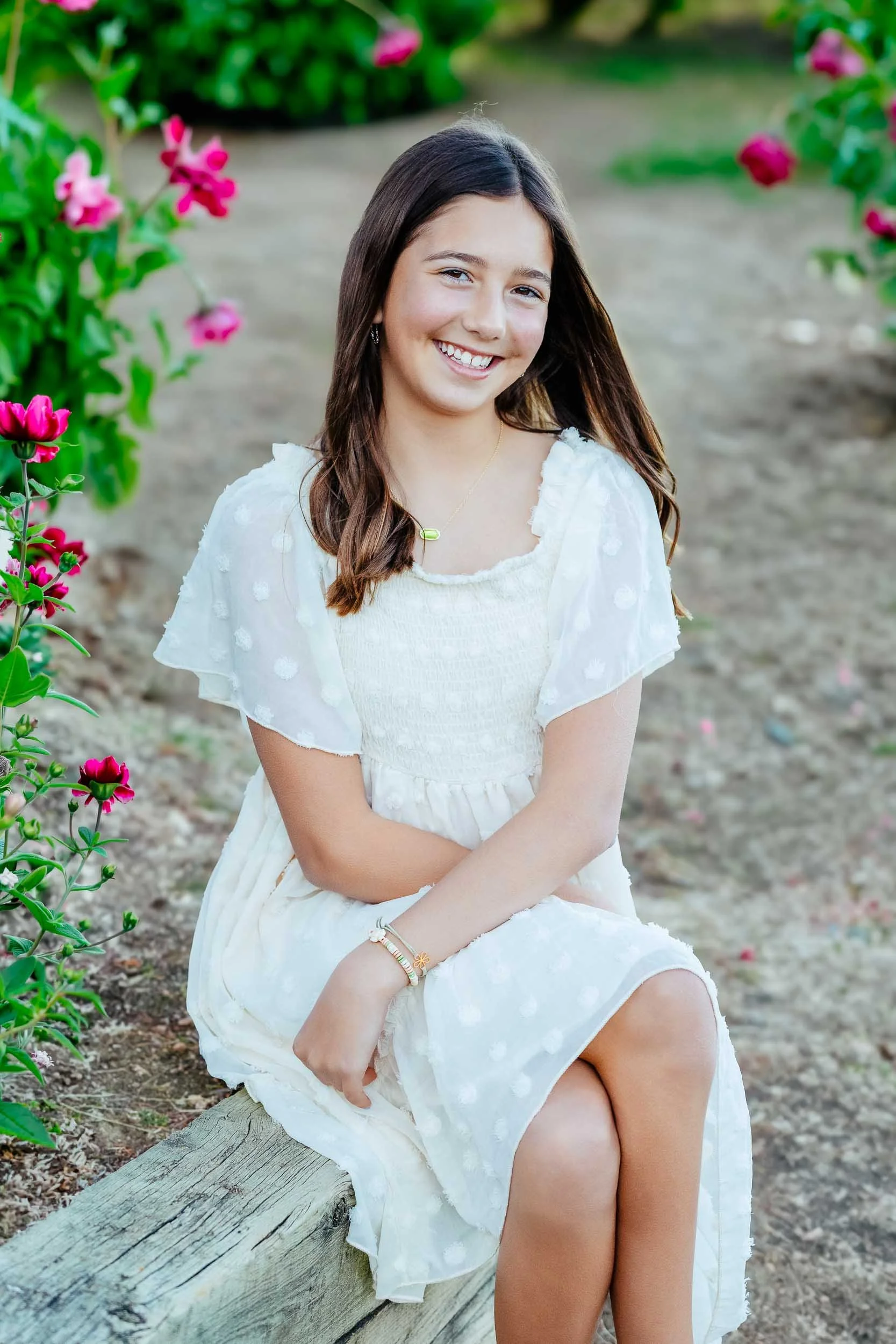 A young girl with brown hair seated on a wooden log in a garden with pink flowers and green foliage, smiling at the camera.