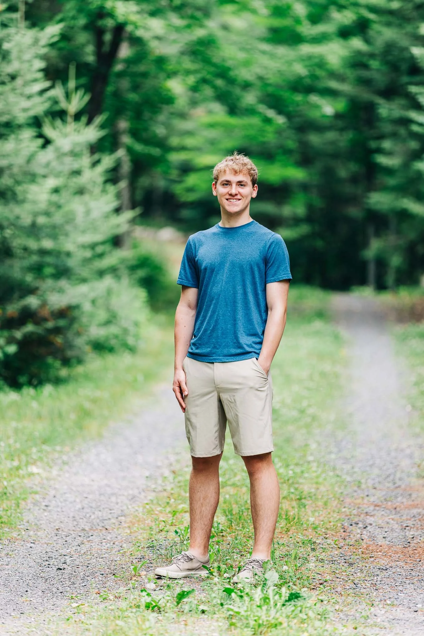 A young man wearing a blue t-shirt and khaki shorts standing on a forest trail.