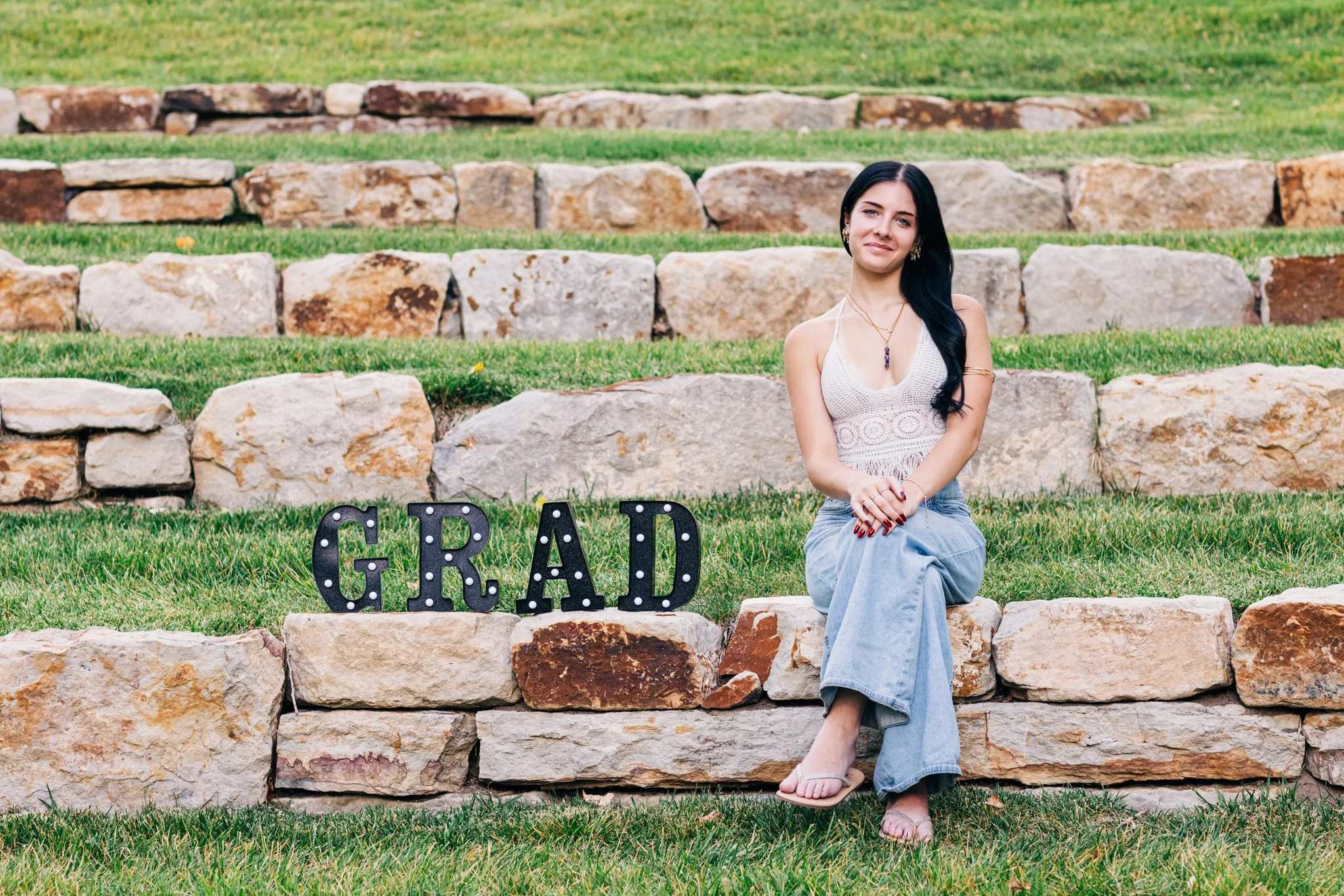 A high school senior with long black hair sitting on a stone wall with a sign that says 'GRAD' beside her, outdoors on grass and tiered stone steps.