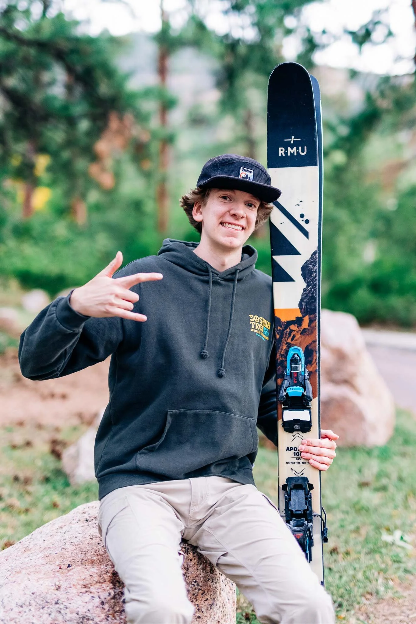 A young man sitting on a rock outdoors holding a ski with a mountain landscape design, wearing a black hoodie and a backward baseball cap, making a hand gesture, with a blurred forest background.