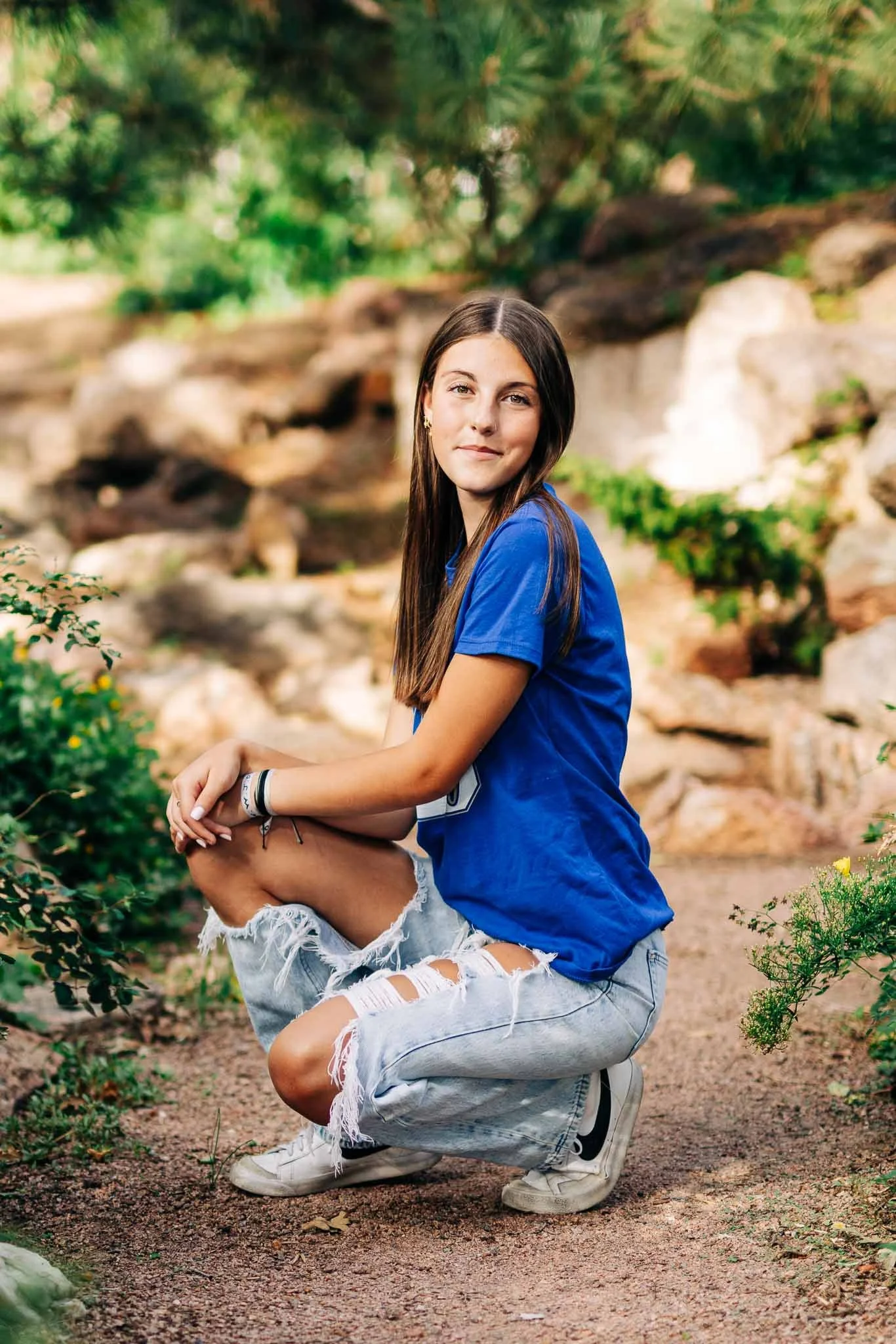 A young woman with long brown hair, wearing a blue t-shirt, ripped jeans, and white sneakers, crouching outdoors on a dirt path surrounded by greenery and rocks.