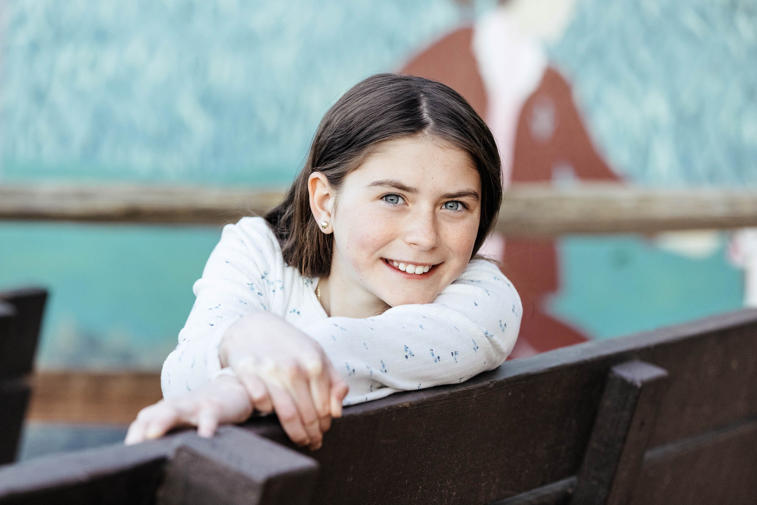 A young girl with short brown hair and blue eyes smiling, leaning on a park bench with her arms crossed, in front of a painted mural.