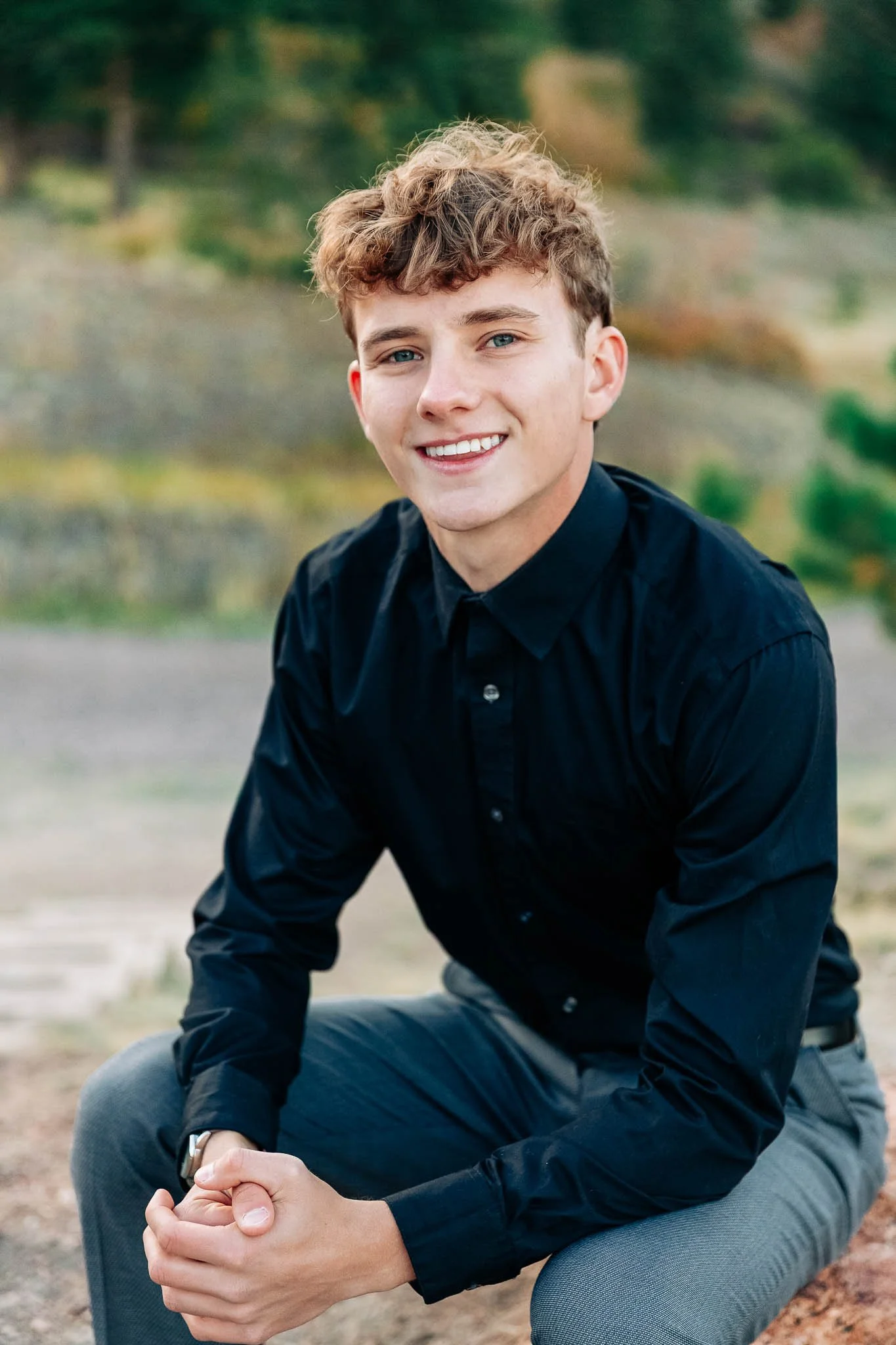 Young man in black shirt and gray pants smiling outdoors with greenery in the background.