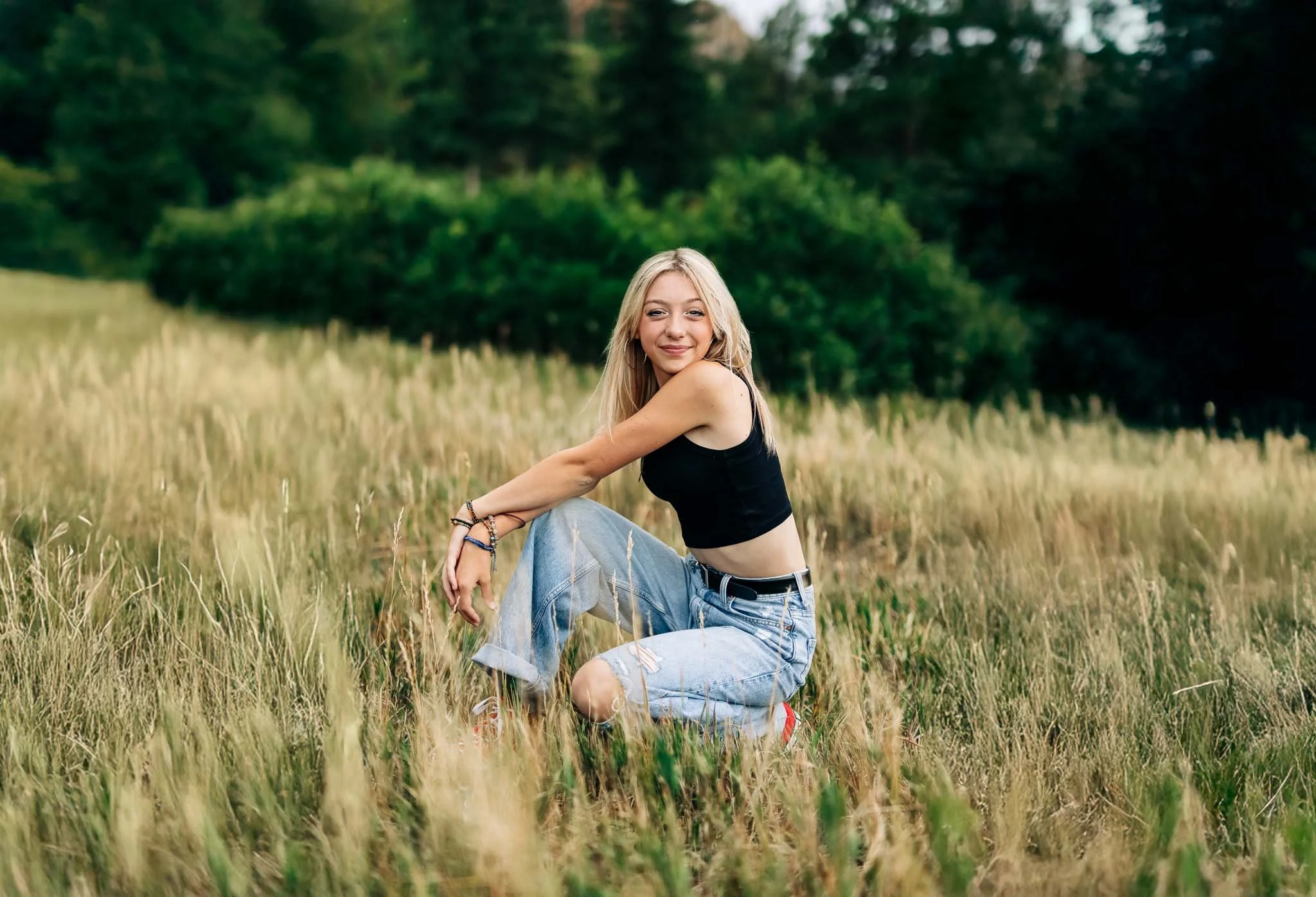 A young woman with blonde hair, wearing a black crop top, ripped jeans, and red shoes, sitting in a grassy field with trees in the background, smiling at the camera.