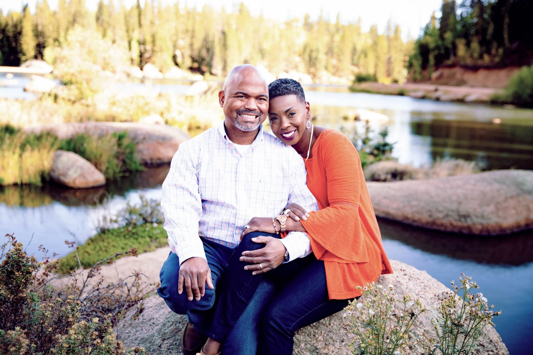 A happy couple sitting on a rock near a lake, surrounded by trees, smiling at the camera.