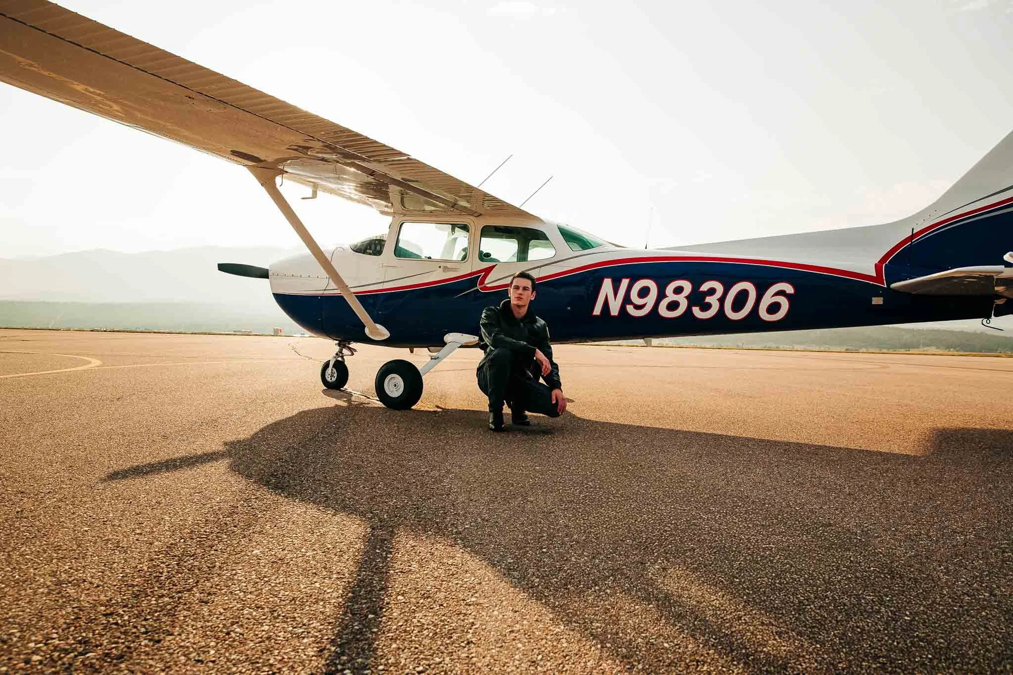 A young man crouching in front of a small private airplane with the tail number N98306 on tarmac, with mountains in the background.