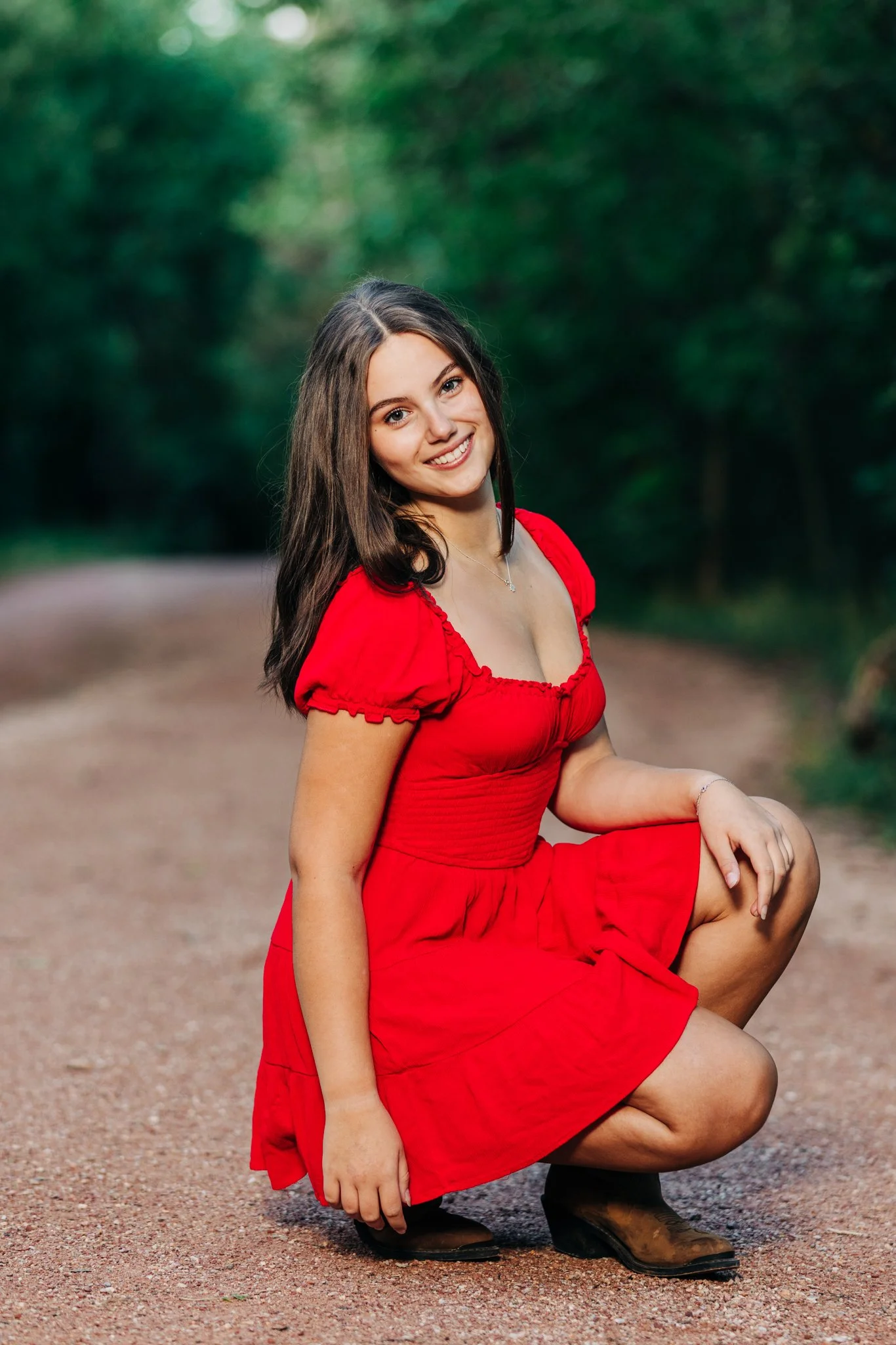 A high school girl with brown hair, wearing a red dress and brown boots, smiling and squatting on a dirt path in a green, wooded outdoor setting.