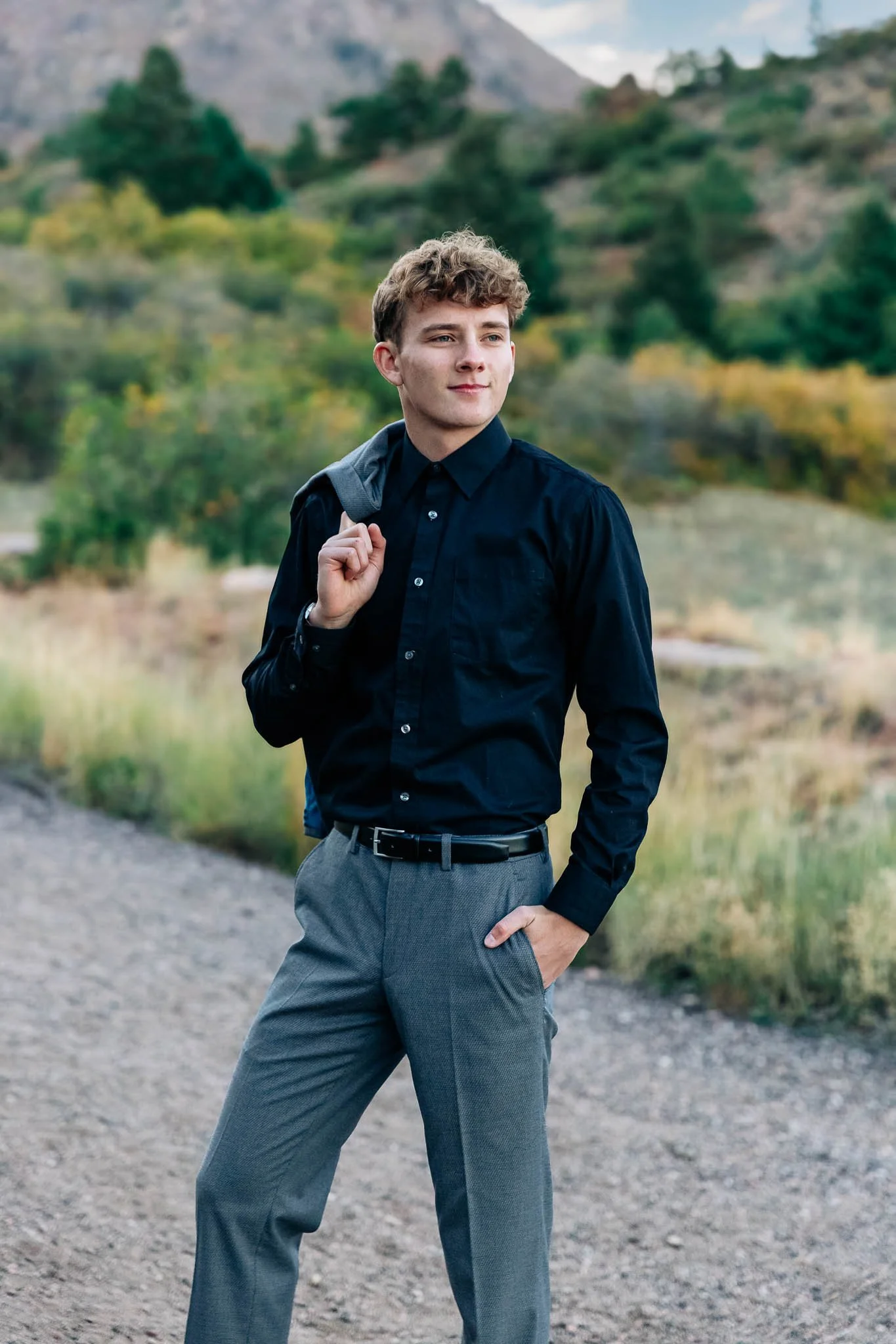 Young man in black shirt and gray pants standing outdoors with a backpack over his shoulder, looking into the distance during daytime with a background of trees and hills.