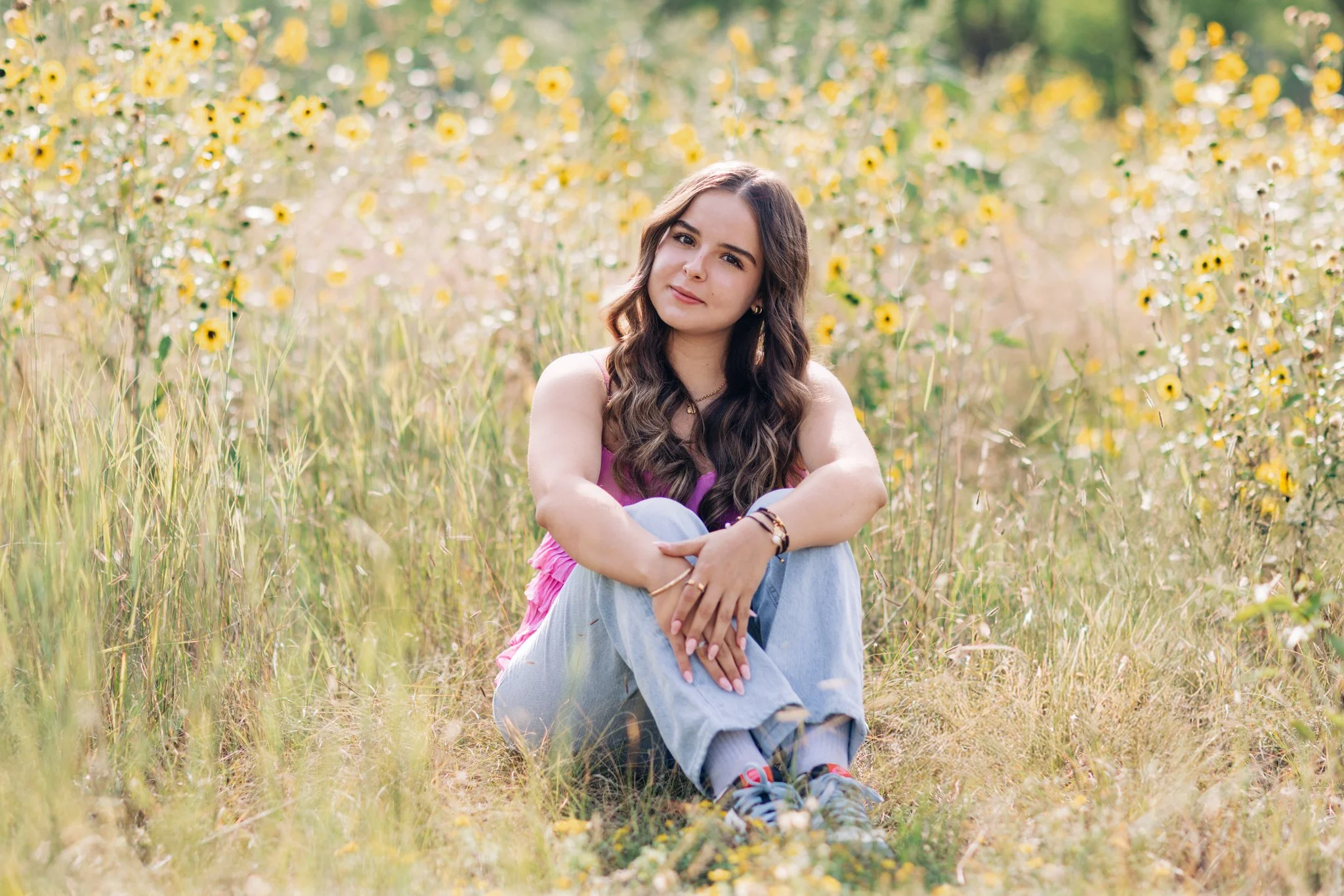 A high school senior with long wavy brown hair sitting in a field of yellow and white flowers. She is wearing a sleeveless pink top, light blue jeans, and colorful sneakers. She has a relaxed expression and is looking at the camera.