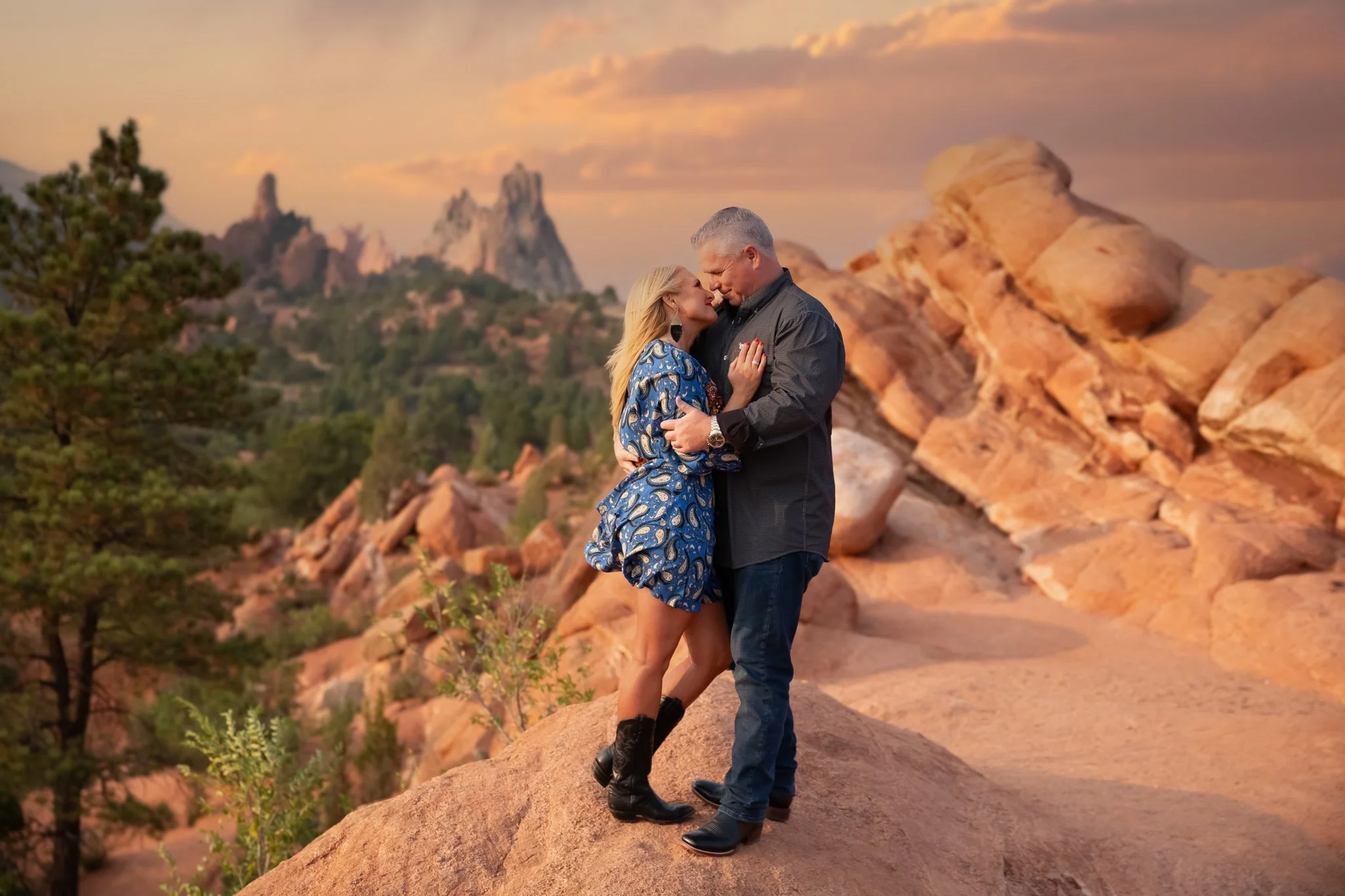 A couple embracing on a rocky outcrop during sunset in a desert landscape with rock formations and trees in the background.