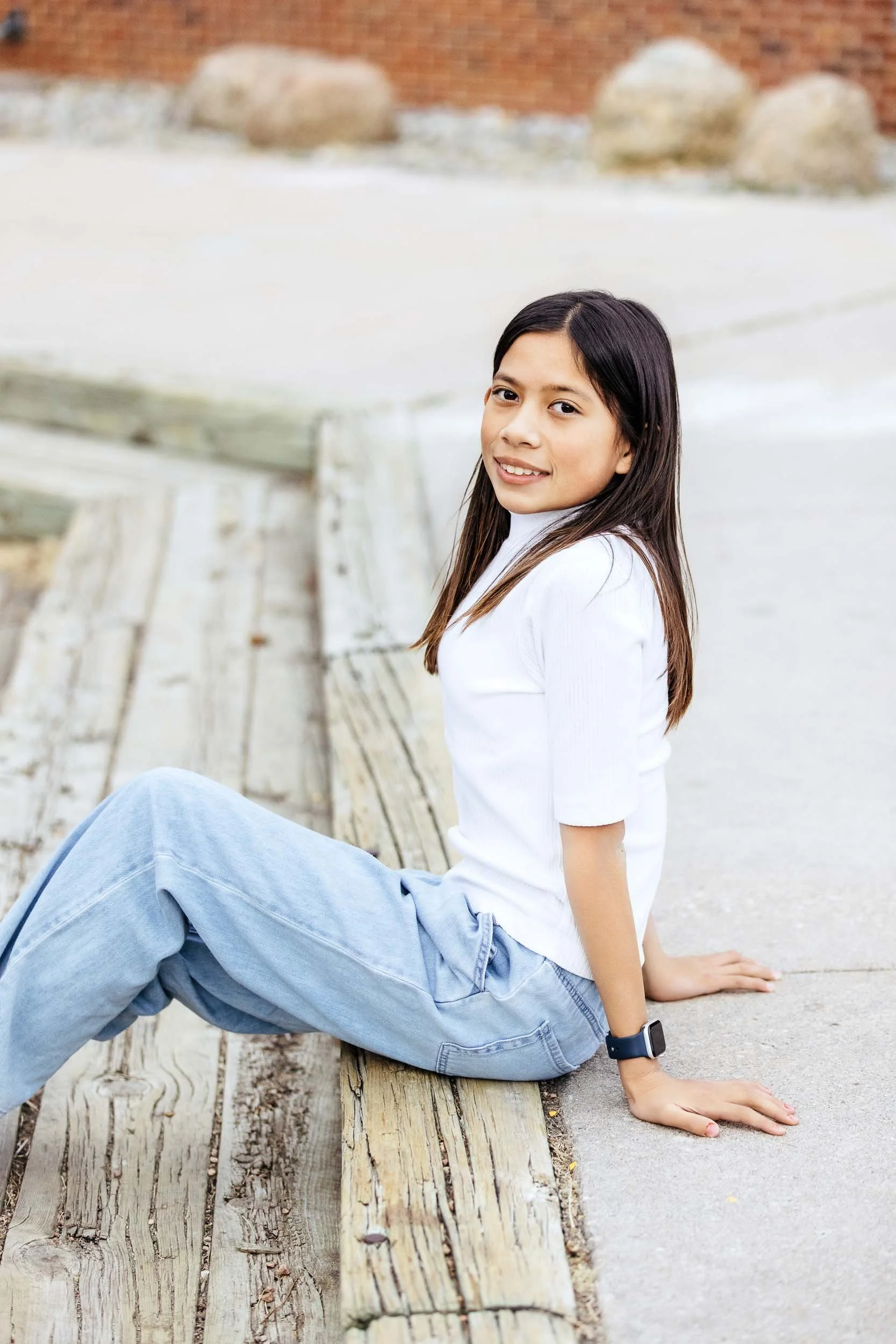 A young girl sitting on a wooden bench outdoors, wearing a white t-shirt, light blue jeans, and a black watch, smiling at the camera.