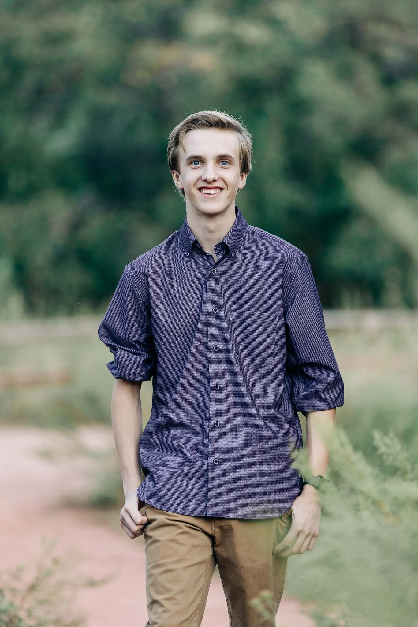 A young man with light brown hair and blue eyes standing outdoors on a dirt path, wearing a purple button-up shirt and tan pants, smiling at the camera with arms relaxed at his sides.