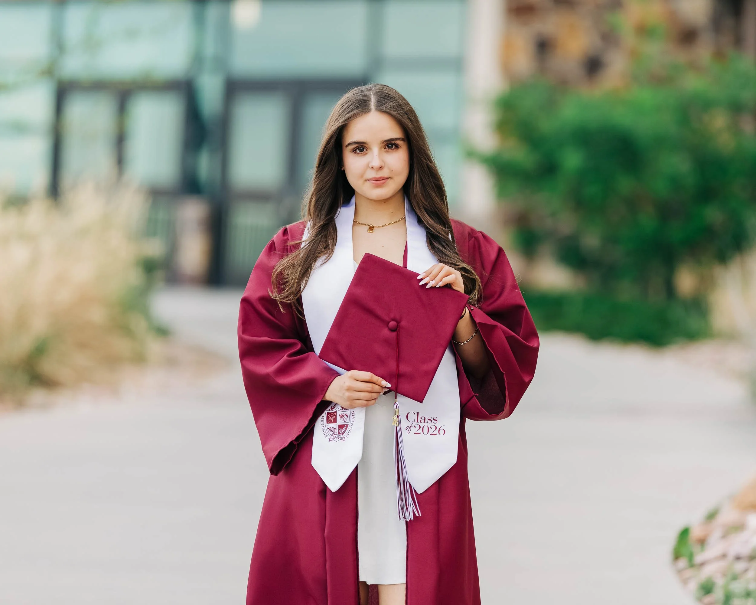 A high school senior in a maroon graduation gown and white dress holding a maroon cap standing outdoors in front of trees and a building.