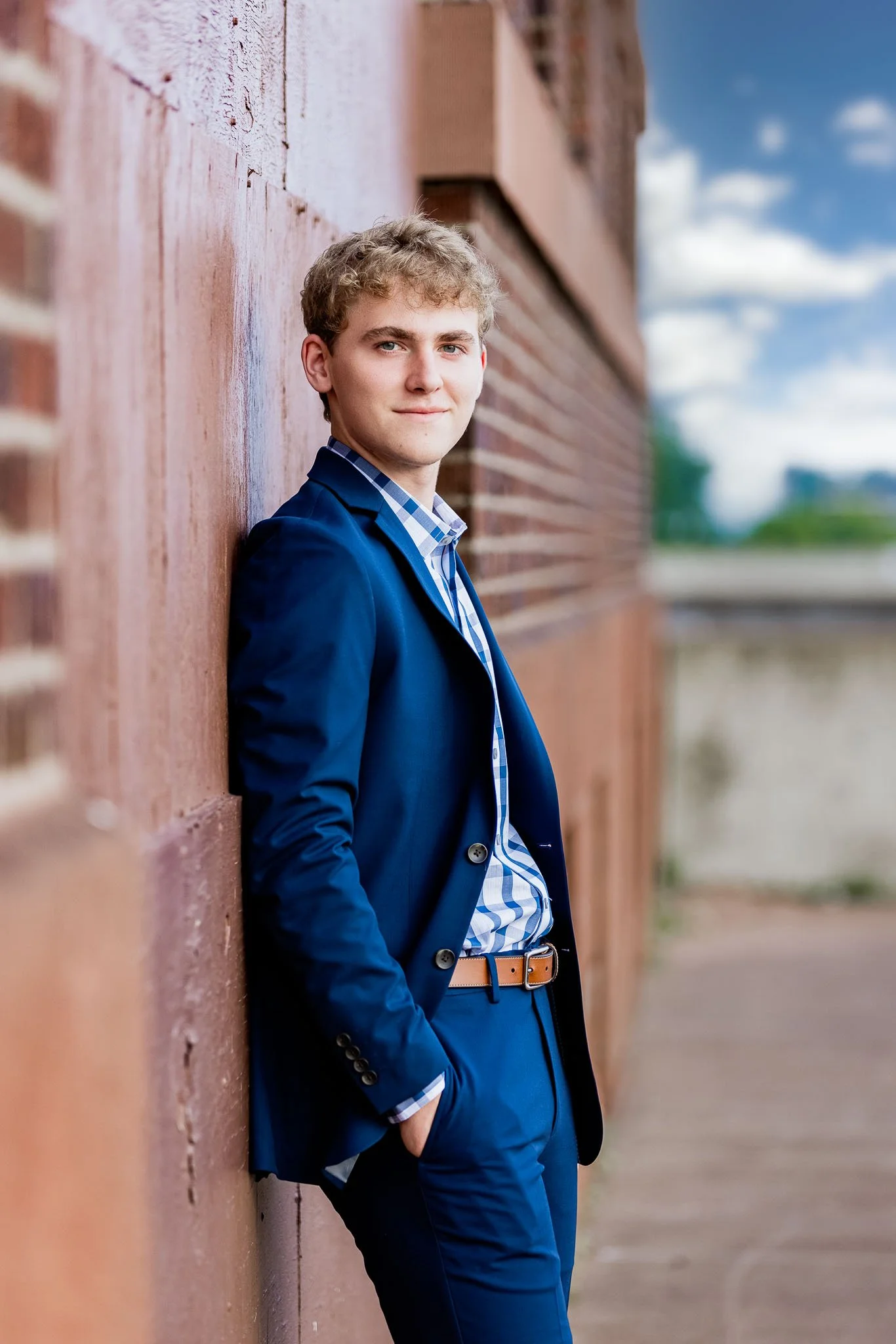 A young man in a blue suit and plaid shirt leaning against a brick wall outdoors, with a blurred background of a partly cloudy sky.