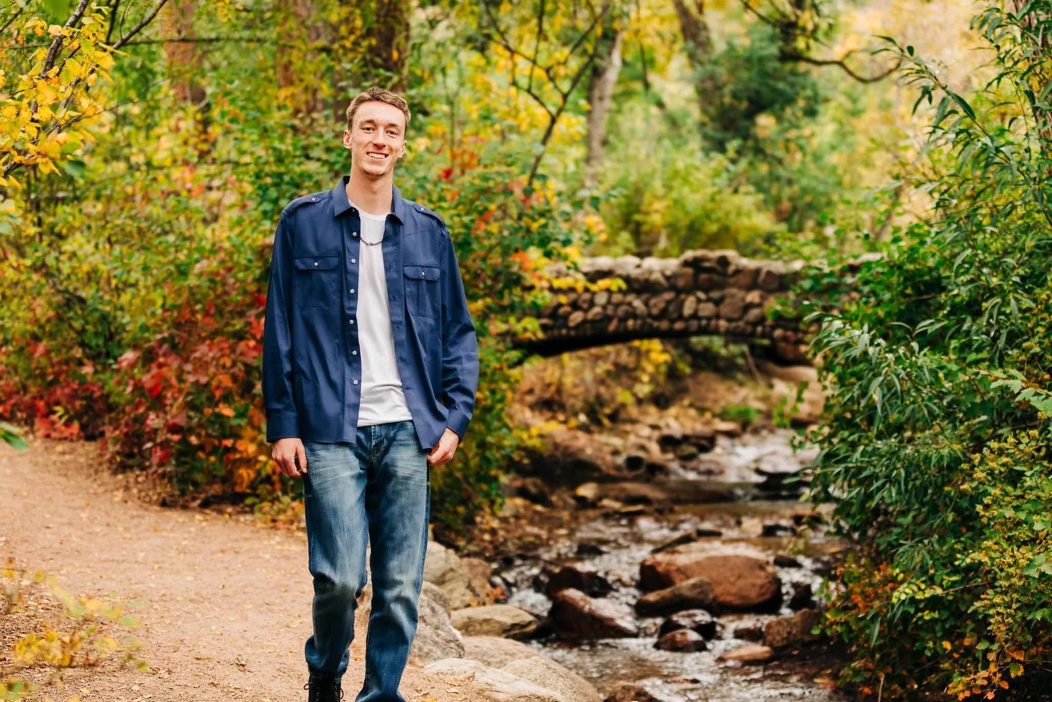 A young man with short hair smiling while walking on a trail through a fall-colored forest near a small creek and stone bridge.